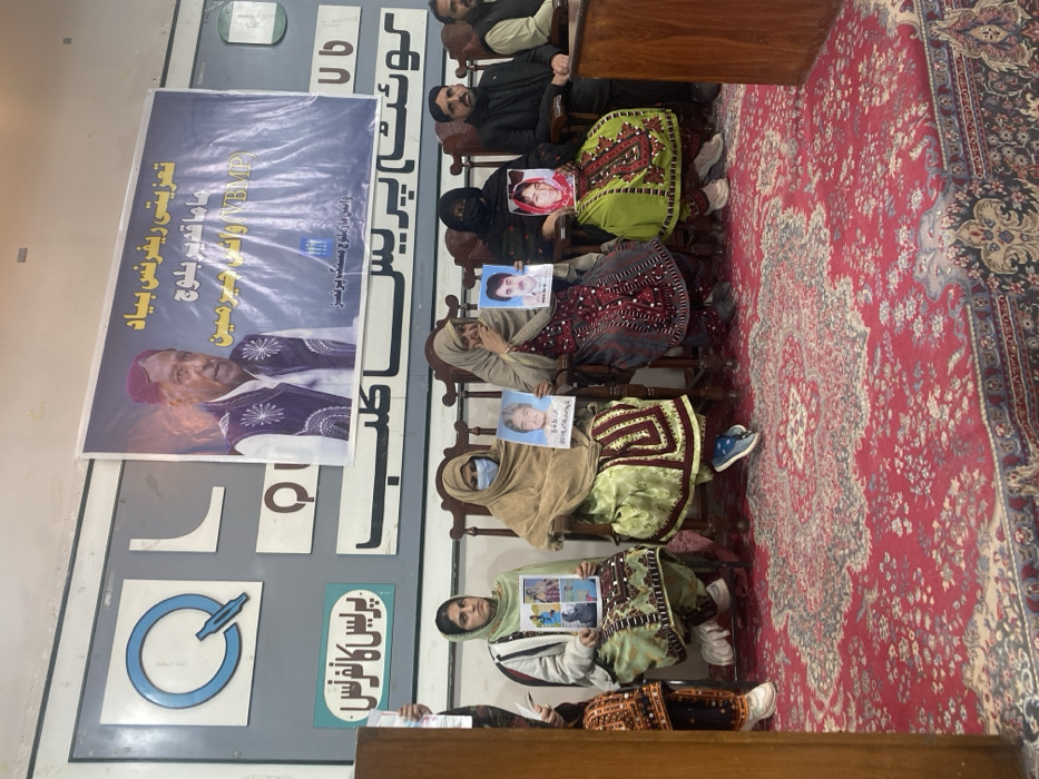 An indoor scene within the Quetta Press Club, located in Quetta City Tehsil, Pakistan, depicts a press conference or gathering. Eight individuals are visible, primarily seated on ornate wooden chairs in the foreground. Five women occupy the left and center, while three men are positioned to the right.
The women are dressed in traditional shalwar kameez attire, with head coverings. The second woman from the left wears a light brown hijab and a medical mask. Each of the five women holds up a framed photograph: the leftmost woman holds a collage of four portraits; the next holds a portrait of a man with Urdu text and the date "2016/04/18"; the third holds a portrait of a man with Urdu text; the fourth holds a portrait of a woman; and the fifth holds another portrait of a man with Urdu text. One woman's hand is raised towards her face, near her nose/mouth, while another's hand is near her chin, possibly indicating distress or contemplation. The men are also dressed in traditional wear, some with jackets.
Behind the seated individuals, a large blue banner is affixed to a light-colored wall. The banner features a portrait of an elderly man with a red cap and a dark embroidered vest, accompanied by Urdu text identifying it as a "Condolence Reference" related to "Mama Qadeer Baloch" and "Voice for Baloch Missing Persons (VBMP)". To the left of the banner, a grey board displays "QUETTA PRESS CLUB" in both English (Q L Q U B) and Urdu characters, along with "پریس کانفرنس" (Press Conference) on a green sign. A wooden podium is partially visible on the far left. The floor is covered by a red, intricately patterned oriental rug. The room is illuminated by overhead artificial lighting.