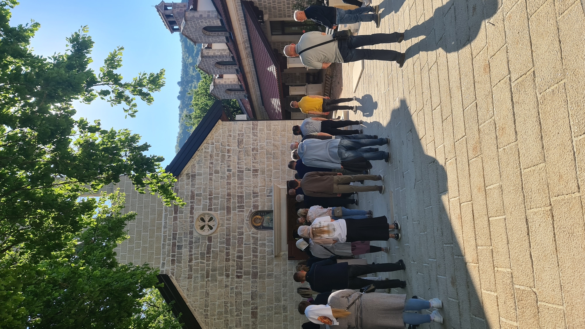 The image shows a crowd of people lined up in front of a stone building. The building appears to be a church. The people are standing on a paved area. A tree is located to the left of the church, and another building is visible in the background. The city is Budva, Montenegro.
