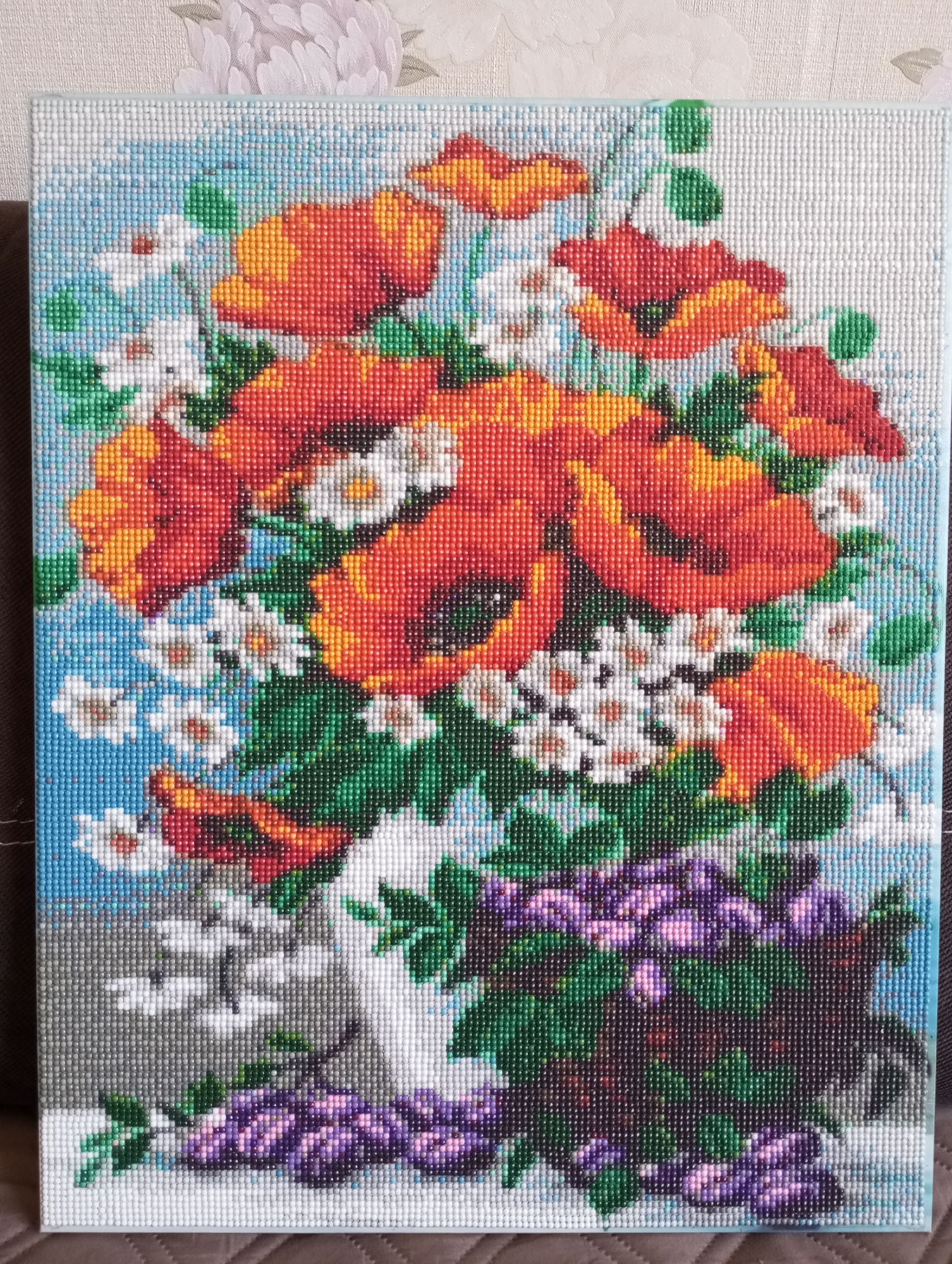 The image depicts a diamond painting of a floral arrangement. The flowers consist primarily of orange and red poppies, along with white daisies and purple flowers arranged in a vase. The background features a mix of blue and white, creating a sky-like effect. The entire image is composed of small, mosaic-like pieces, indicative of a diamond painting technique. The location of Буренка, Russia, is not depicted in the image.