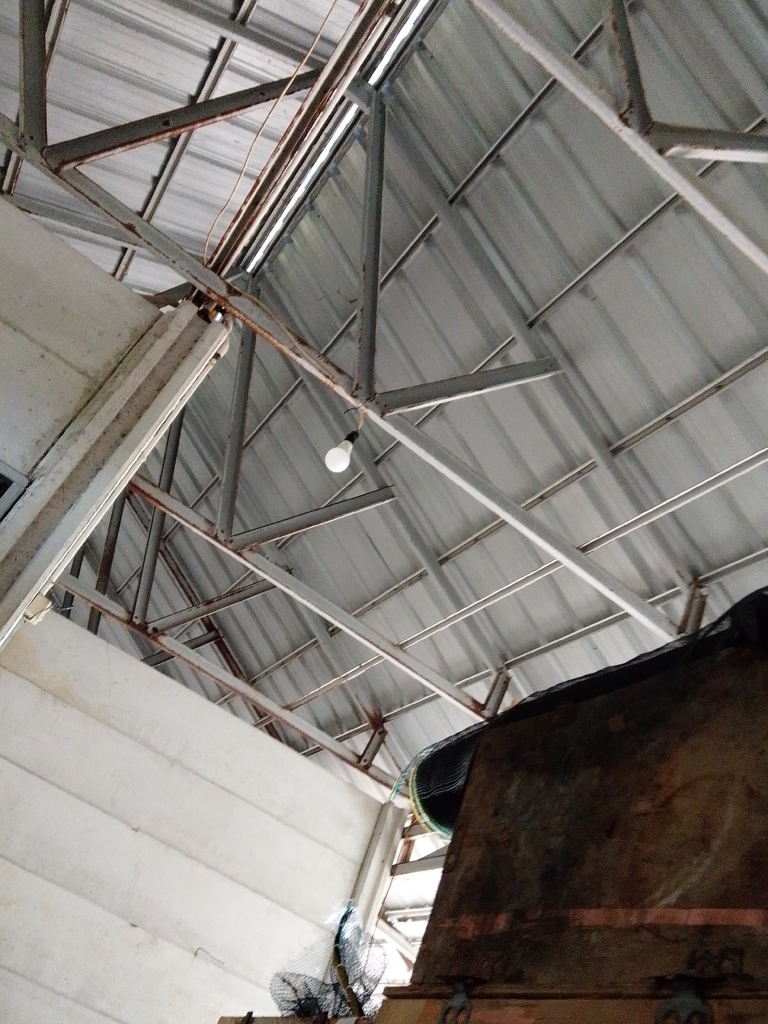 The image, taken indoors in Bukit Ketok, Indonesia, depicts a low-angle view of a ceiling structure. The roof is constructed of corrugated metal panels supported by a framework of metal beams and supports, some of which exhibit rust. A single lightbulb hangs from the metal framework. A white wall with horizontal panels is visible in the lower-left corner. A large, dark, textured object occupies the lower-right corner, partially obscuring the view. A fishing net is also visible. The scene is dimly lit.