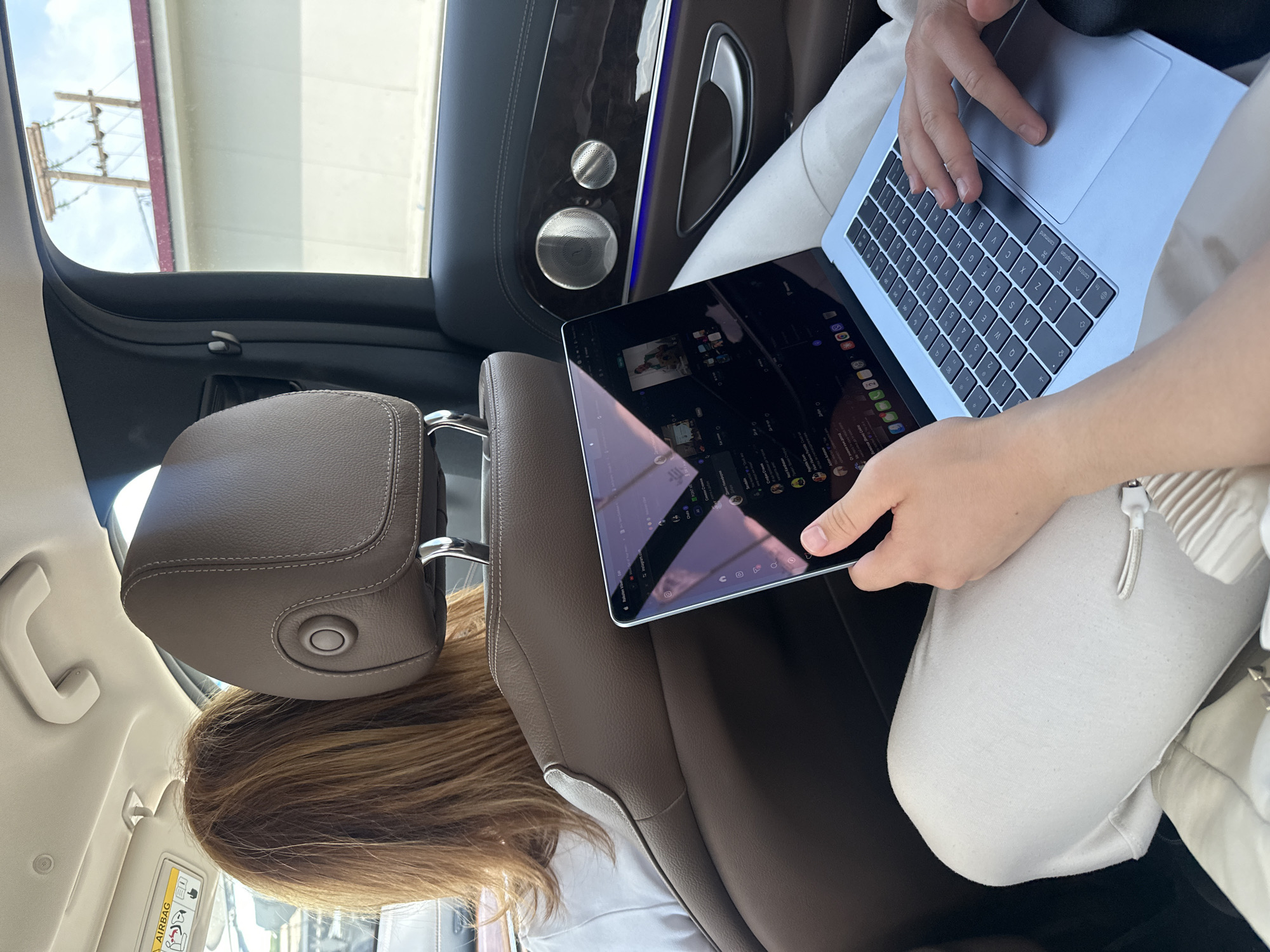 The image captures a daytime scene inside the rear passenger area of a car, viewed as if the camera was rotated 90 degrees clockwise. When viewed upright, it shows two individuals within the vehicle.
In the foreground, a person is seated, with only their hands, lap, and lower torso visible. They are engaged with two electronic devices: an iPad held in their left hand, displaying a digital interface that appears to be a web browser with various icons and tabs; and a light-colored laptop resting on their lap, its QWERTY keyboard clearly visible with standard keys like "control," "option," and "command."
In front of this person, the back of another individual's head and shoulders are seen, indicating they are occupying the front passenger seat. This person has long, light brown hair. The car's interior features luxurious details including brown leather seats with prominent white stitching on the headrest and seat, a dark wood trim on the passenger door, chrome door handles, and two circular speaker grilles, suggesting a premium sound system. A line of blue ambient lighting runs along the door trim.
Through the car's windows, bright daylight is evident, along with a glimpse of the sky and a building or structure. An old wooden utility pole with multiple crossarms and wires stands outside, hinting at a rural or semi-urban environment. A white sun visor on the passenger side displays "AIRBAG" warning text and symbols. The scene depicts activity within a modern vehicle, set in Δημοτική Ενότητα Προμαχώνος, Greece.