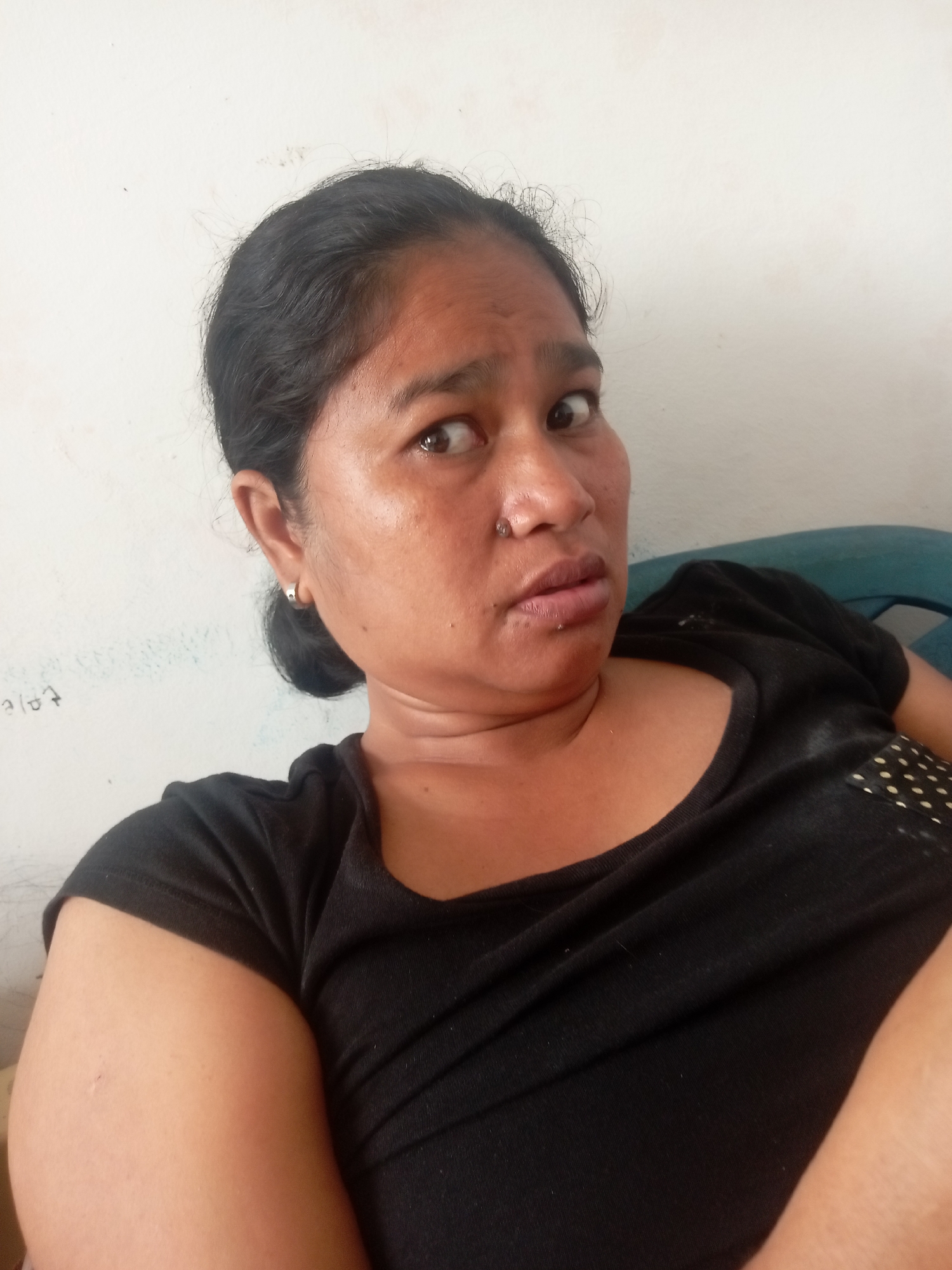 A medium shot captures a woman of Asian descent from the chest up, looking directly at the camera with a slightly surprised or questioning expression. She has dark hair pulled back, exposing her forehead and ears. A small silver hoop earring is visible in her left ear, and a silver stud is in her nostril. She is wearing a black t-shirt that appears slightly wrinkled. The background consists of a plain white wall, with some faint scribbles or markings on it. To the right of the woman, a portion of a teal plastic chair is visible. The lighting suggests an indoor setting, possibly a home or a simple office space. The overall impression is a casual, unposed selfie taken during the daytime. The image is likely taken in Batam, Indonesia, given the inferred location of the subject.