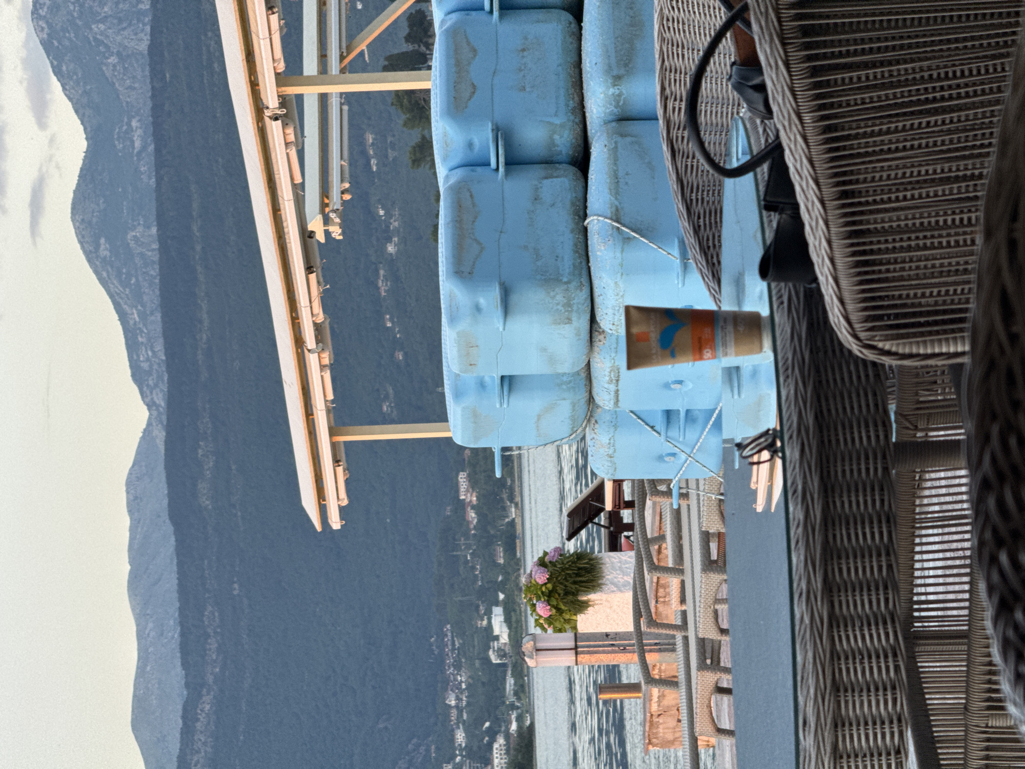 The image, taken in Bogišići, Montenegro, shows a waterfront view from what appears to be a balcony or patio. In the foreground, there is a wicker chair and a stack of light blue, plastic flotation devices. An opened tube of cream is placed on the blue platform. In the middle ground, there's a flower pot with purple flowers. Beyond that lies the sea, partially obscured by buildings along the shore, including the town of Bogišići. In the background, there are mountains. The sky is overcast.