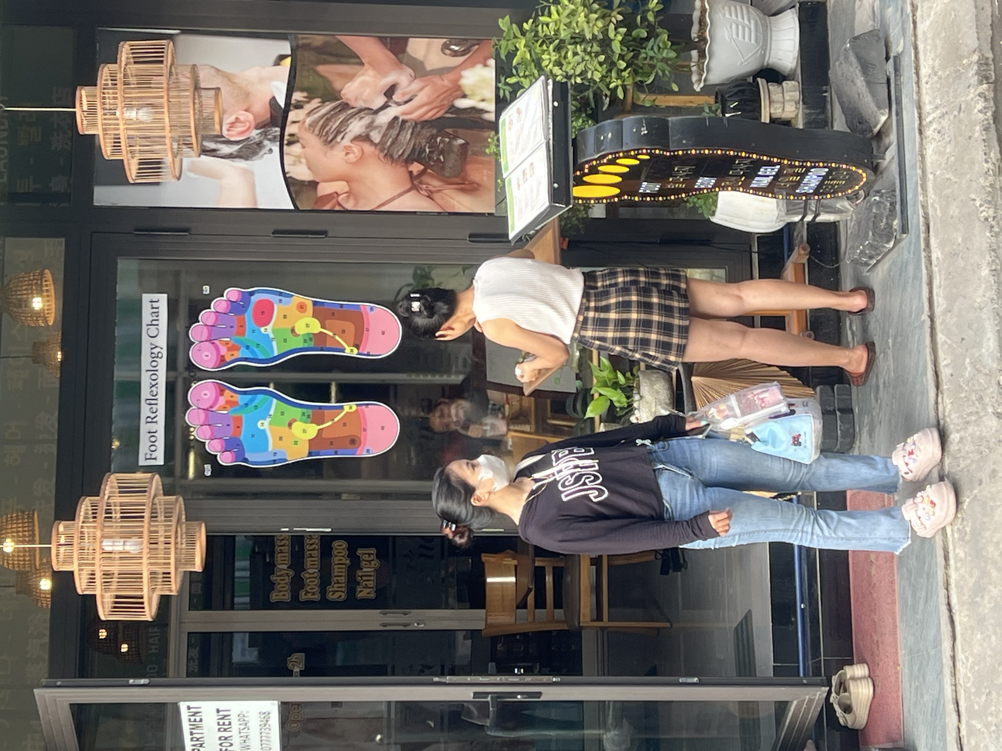 Two Vietnamese women stand outside a salon in Da Nang, Vietnam. The woman on the left, wearing a black hoodie and jeans, has a white face mask and a necklace. She holds a light blue shopping bag and a clear plastic bag filled with pink items. The woman on the right, wearing a white sleeveless top and a plaid skirt, faces away from the camera and appears to be interacting with a menu board. The salon entrance is decorated with a colorful foot reflexology chart and a photograph of someone receiving a hair wash. Other elements visible include a wooden chair inside the salon, several potted plants, and a unique foot-shaped sign with illuminated text. The setting suggests a commercial street scene.