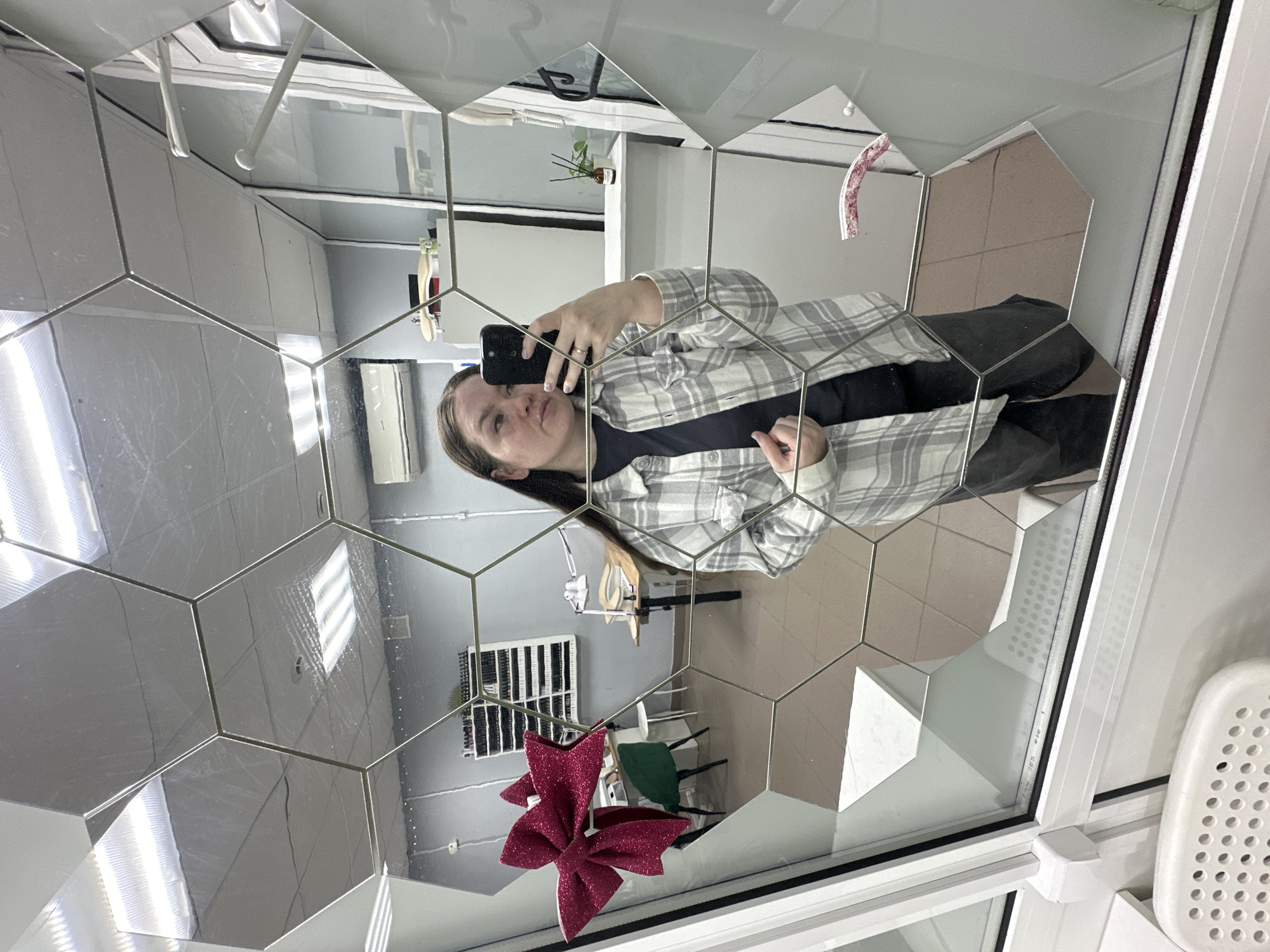 This image, captured in the city of Chaikovsky, Russia, depicts a woman taking a selfie in a mirror constructed from hexagonal tiles. The woman has long brown hair and is dressed in a light-colored plaid shirt over a dark t-shirt and dark pants. She is holding a dark-colored smartphone, which partially obscures her face, in her right hand. A ring is visible on her left ring finger, and her fingernails are painted a light hue. The hexagonal mirror tiles, some exhibiting smudges, are bordered by white window or door frames on the left and bottom. In the immediate foreground, a white perforated stool is partially visible below the mirror.
The mirror's reflections reveal the interior of a room with a white drop ceiling that features rectangular fluorescent light panels and a smoke detector. The walls are painted a light grey. On the left side of the reflection, a white shelving unit filled with numerous small bottles, consistent with nail polish or gel colors, is situated against the far wall. Adjacent to this, a light-colored desk with a white task lamp and tools is visible, accompanied by a green chair and a white chair. A red glittery bow decoration is affixed to the mirror or its frame on the far left. On the right side of the reflection, an air conditioning unit is mounted high on the wall, and a white counter or shelf displays a small potted plant and a diffuser bottle. Portions of a white door frame and an intercom unit are also reflected. The floor, visible in the lower mirror tiles, is tiled with light beige or brown squares. The overall environment indicates a professional setting, likely a beauty salon or studio.