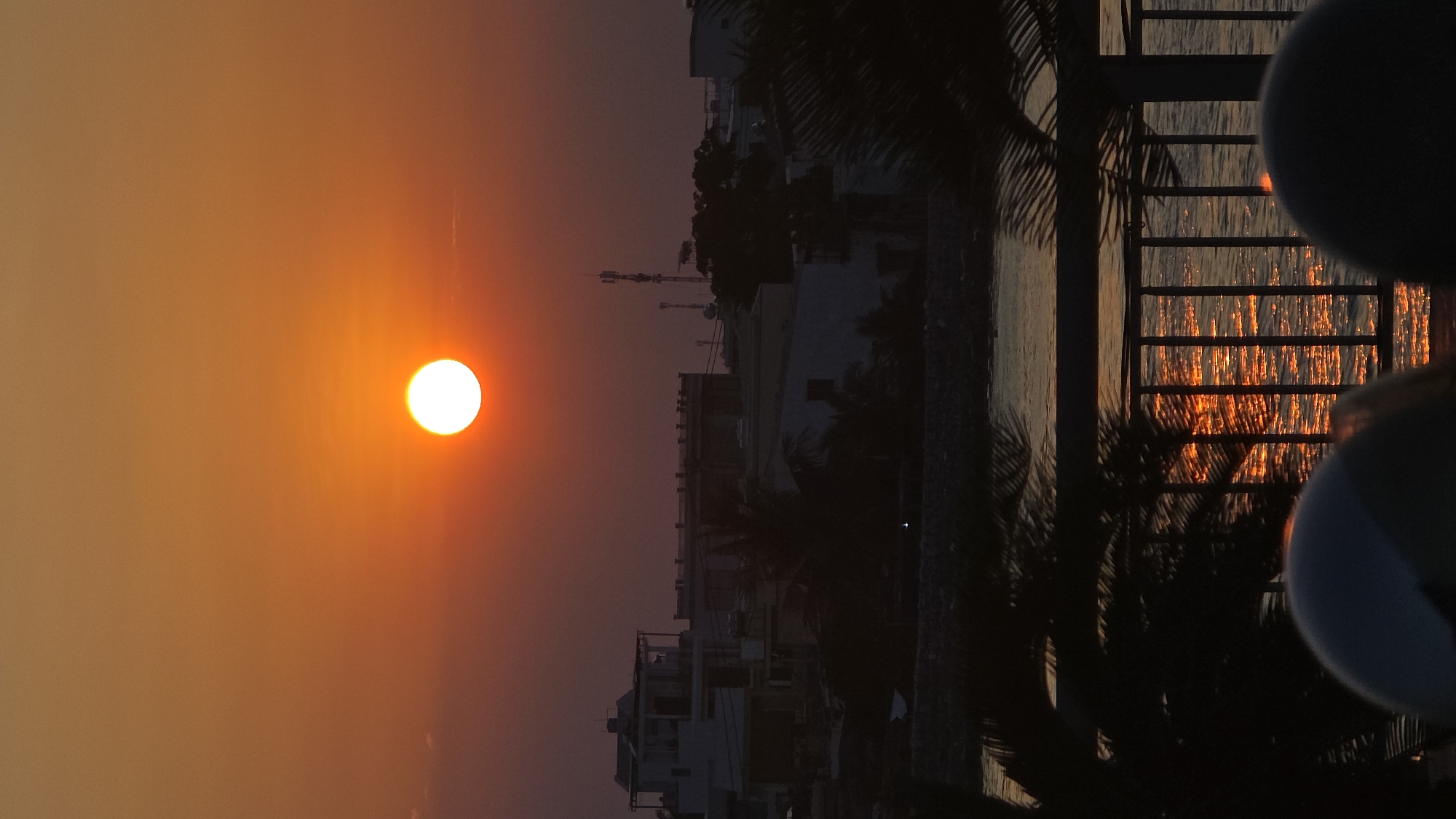 An outdoor scene captured during sunset in Nha Trang, Vietnam, presents a coastal urban landscape. The sky is dominated by an orange-red gradient, with the bright, white-yellow orb of the sun positioned centrally in the upper half of the frame. Below the horizon, a silhouetted urban environment features multiple low to medium-rise structures, including a prominent white building with a flat rooftop on the left. Several communication towers are discernible against the sky on the right side of the skyline. Interspersed palm trees and other dark foliage are visible within the cityscape. A body of water occupies the mid-ground, reflecting the sun's light in vertical streaks of orange and yellow. A dark, possibly rocky or constructed, shoreline separates the water from the urban structures. In the immediate foreground, a dark metal railing or fence extends horizontally, partially obscured by the dark silhouettes of palm fronds on the left and right edges. Two rounded, dark objects, consistent with the tops of helmets or human heads, are partially visible at the bottom of the frame, suggesting the presence of observers. No discernible actions or interactions are visible.