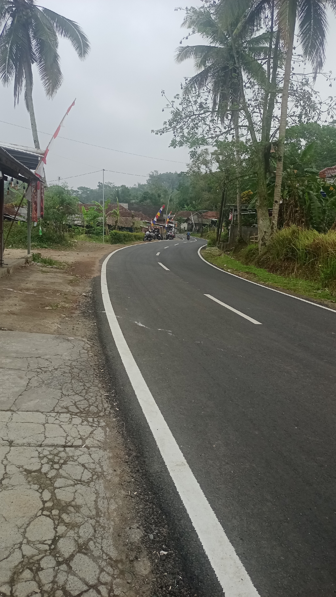 The image shows a paved road curving to the left through a rural area in Cogreg, Indonesia. The road has a white line marking its edge and another white line dividing the lanes. To the left of the road, there is a cracked concrete sidewalk and a dirt area. A few buildings with tin roofs are visible in the background, along with trees and vegetation. Several motorcycles are parked near some of the buildings, and a single person on a motorcycle is riding on the road. Indonesian flags are displayed on poles near the buildings. The sky is overcast.