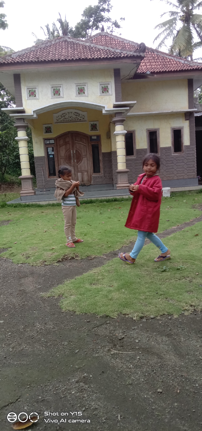 A daytime outdoor scene in Ranugedang, Indonesia, features two young girls on a grassy area in front of a two-story building. The girl on the left stands facing forward, wearing a light-colored striped shirt, light brown pants, and red sandals. She is wrapped in a patterned brown and beige shawl, holding it with both hands in front of her midsection. The girl on the right is in motion, stepping from right to left, wearing a red long-sleeved jacket, light blue pants, and purple sandals. Her hands are raised slightly, positioned near her chest.
The background structure is a building with a light yellow stucco facade, accented by darker brown or grey elements on its pillars and lower walls. It has a reddish-brown tiled roof with a distinctive curved eave and some ornamental features on the ridge. The central entrance consists of double wooden doors with circular carved details, surmounted by a carved white archway. Four recessed square panels with red and green floral designs are visible on the upper facade. The right side of the building shows dark, rectangular openings, possibly unglazed windows. The ground consists of green grass interspersed with dark, exposed soil paths. Tropical trees, including visible palm fronds, are present in the background.