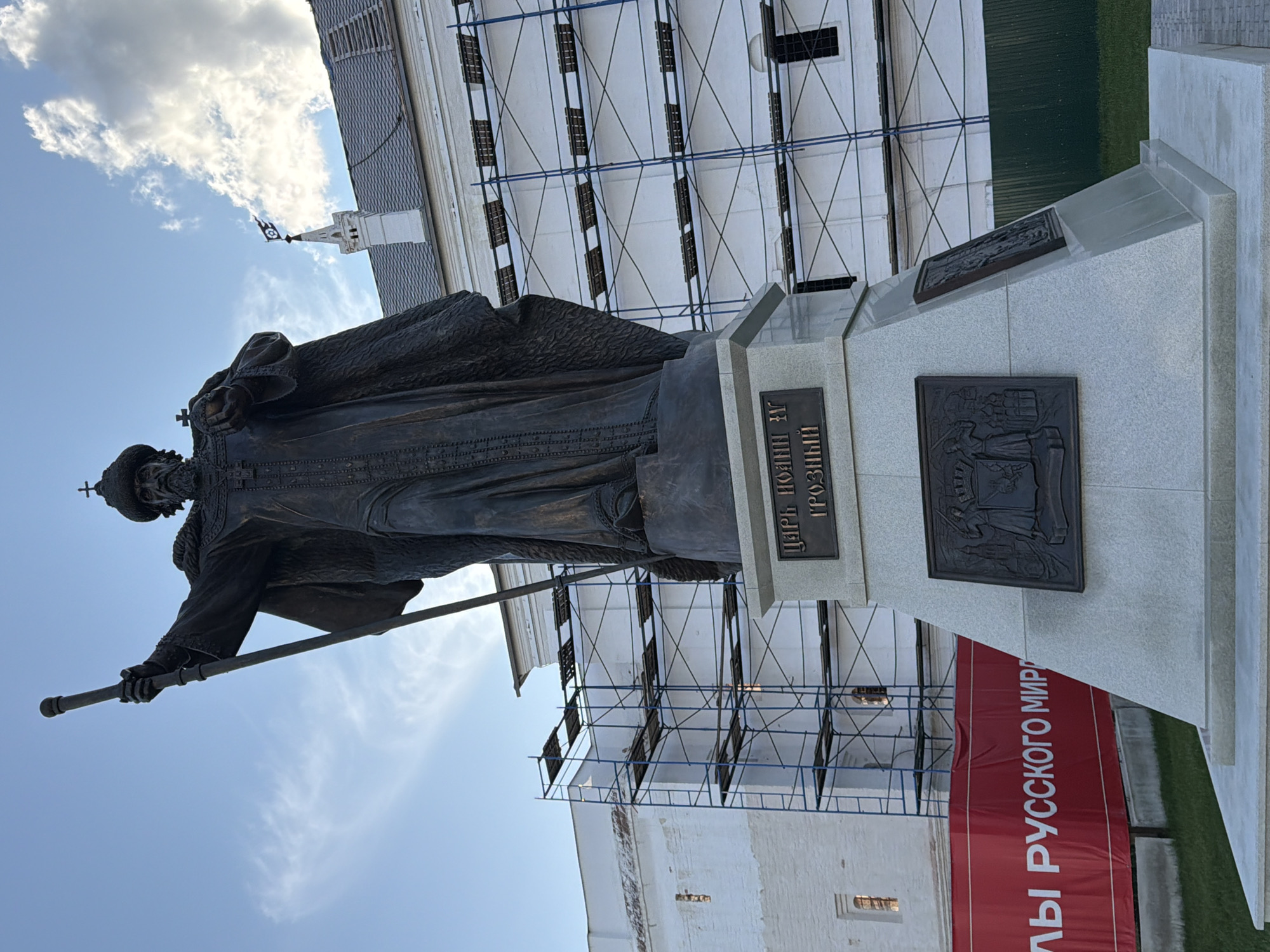 The image shows a bronze statue of Ivan the Terrible in Vologda, Russia. The statue is located in front of the Vologda Kremlin, which is currently undergoing renovations and is partially covered by scaffolding. The statue depicts Ivan IV as a stern, imposing figure, holding a scepter in his right hand and a cross in his left. He is wearing a regal robe and a crown. The base of the statue is inscribed with the words "ЦАРЬ ИОАНН IV ГРОЗНЫЙ" (Tsar Ivan IV the Terrible). In the background, a red banner with white lettering is visible, partially obscured by the statue. The sky is partly cloudy.