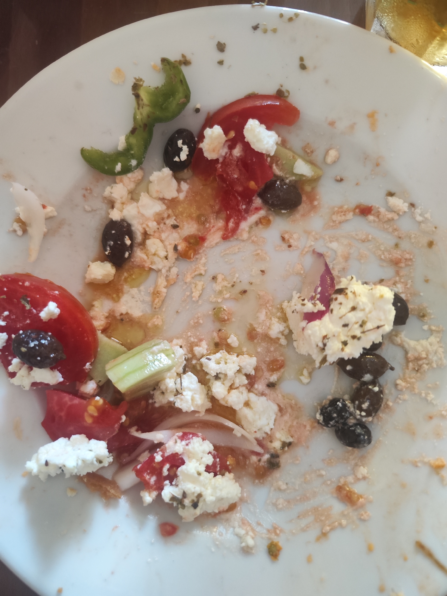 The image depicts the remnants of a Greek salad on a white plate, likely at a restaurant in Melissourgeio, Greece. The plate is sparsely populated with scattered ingredients: chunks of feta cheese, sliced tomatoes, black olives, and pieces of green bell pepper and cucumber. Traces of dressing and other food particles are spread across the plate, suggesting the meal has been consumed. The background, out of focus, appears to be a wooden table, and a glass of a light-colored beverage is partially visible at the top right. The lighting is natural, creating shadows and highlighting the textures of the food residue. There are no people or visible actions occurring in the scene.