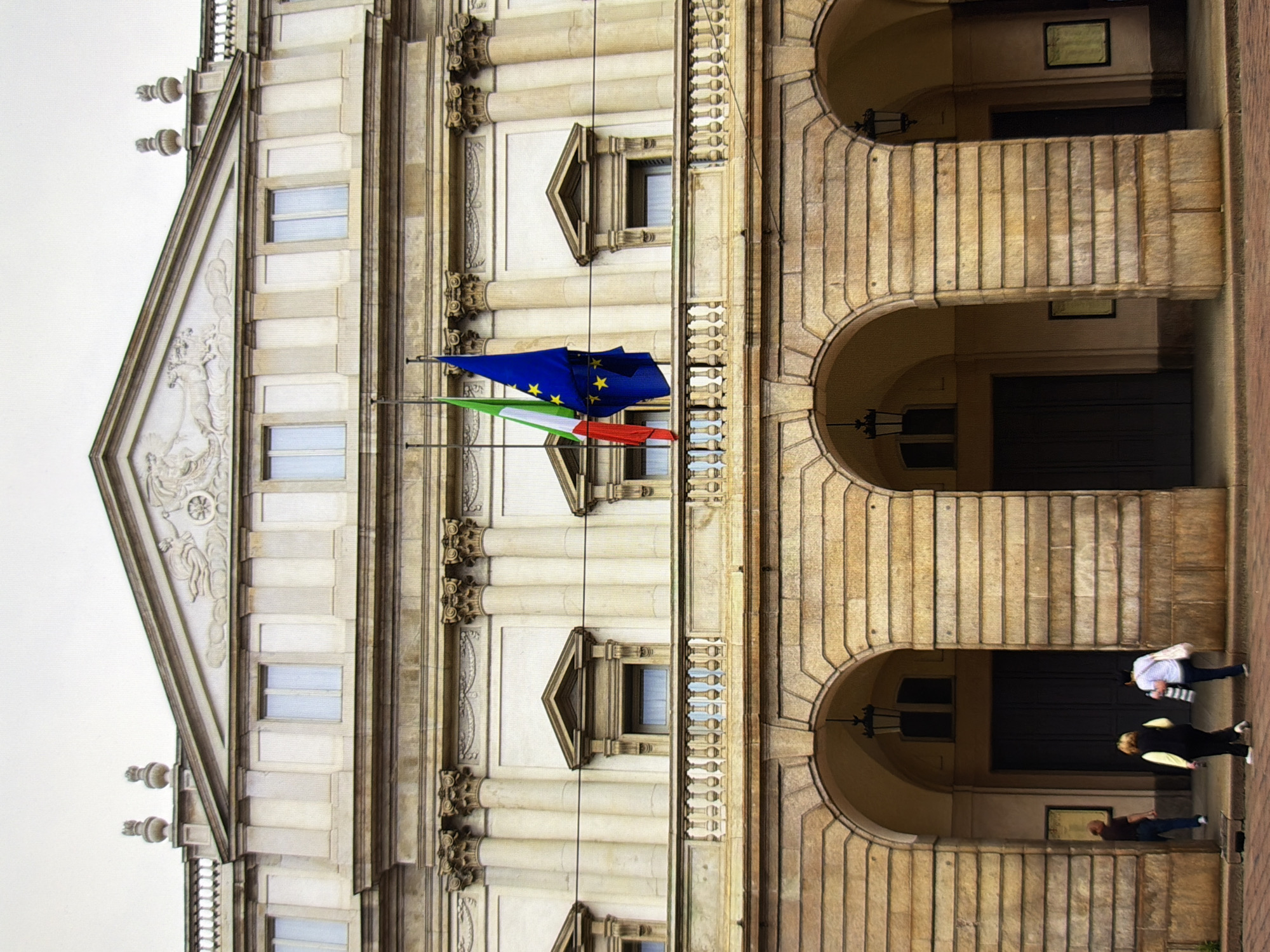 The image shows a grand neoclassical building, possibly a theater or opera house, with a light-colored stone facade. The building has multiple levels, adorned with columns and decorative architectural elements. Two flags, one green, white, and red (likely the Italian flag) and a blue flag with yellow stars (the European Union flag), are visible on poles extending from the building. In the foreground, three people are seen walking towards the building's arched entrance. One person, wearing a dark shirt and blue jeans, is entering the building on the left. Two other individuals, one wearing a black vest over a yellow top and the other in a white top and blue jeans, are walking into the building from the center. The ground in front of the building appears to be paved with brick. There are no identifiable landmarks or specific details to indicate the location is in Kyiv, Ukraine.