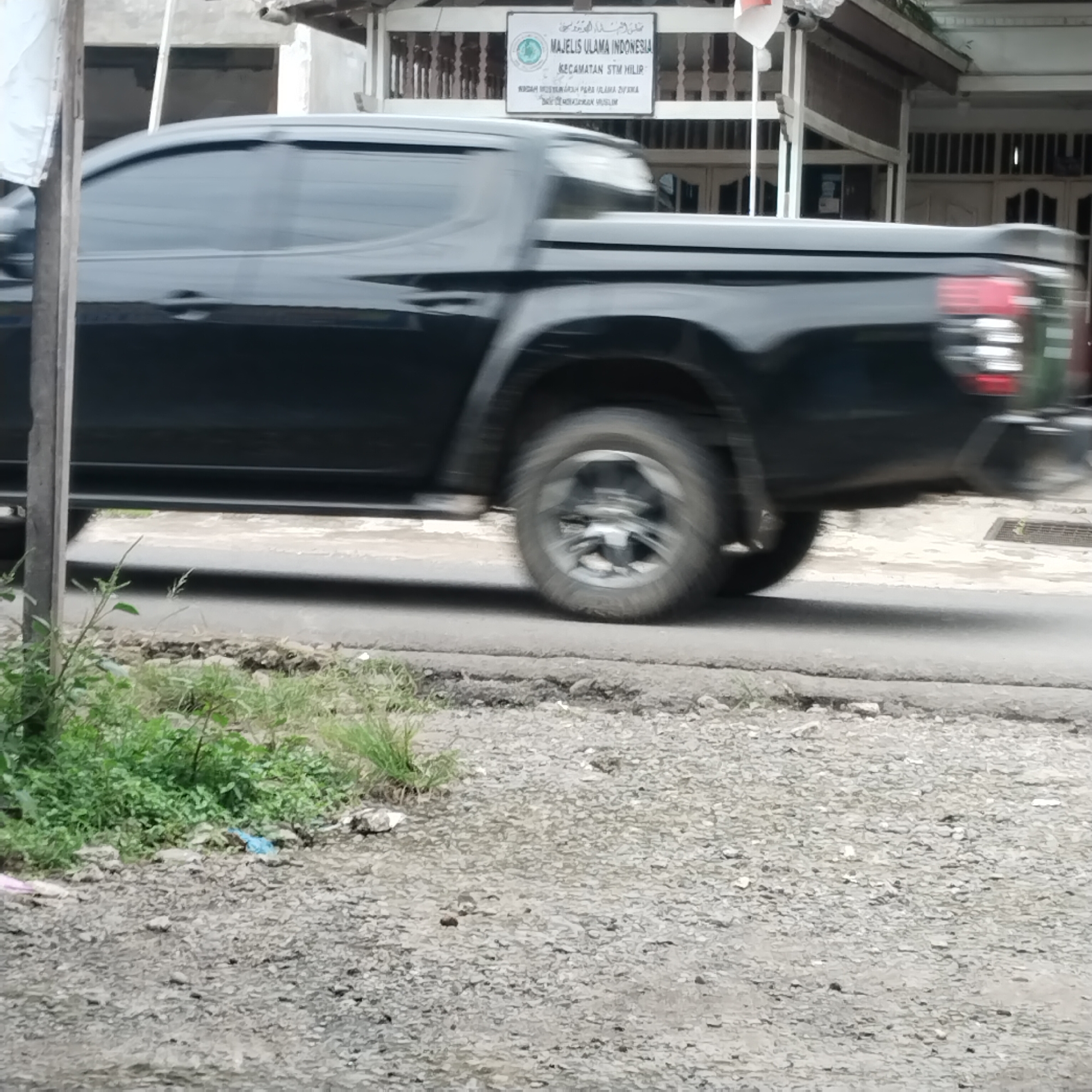 The image shows a black pickup truck driving on a road. The truck is in motion, and its wheels are blurred. The truck is black and has tinted windows. In the background, there is a building with a sign that says "MAJELIS ULAMA INDONESIA". The building appears to be a mosque or a religious institution. There are also some trees and grass visible in the background. The ground in front of the building is covered with gravel. The image appears to have been taken in the city of Talun Kenas, Indonesia.