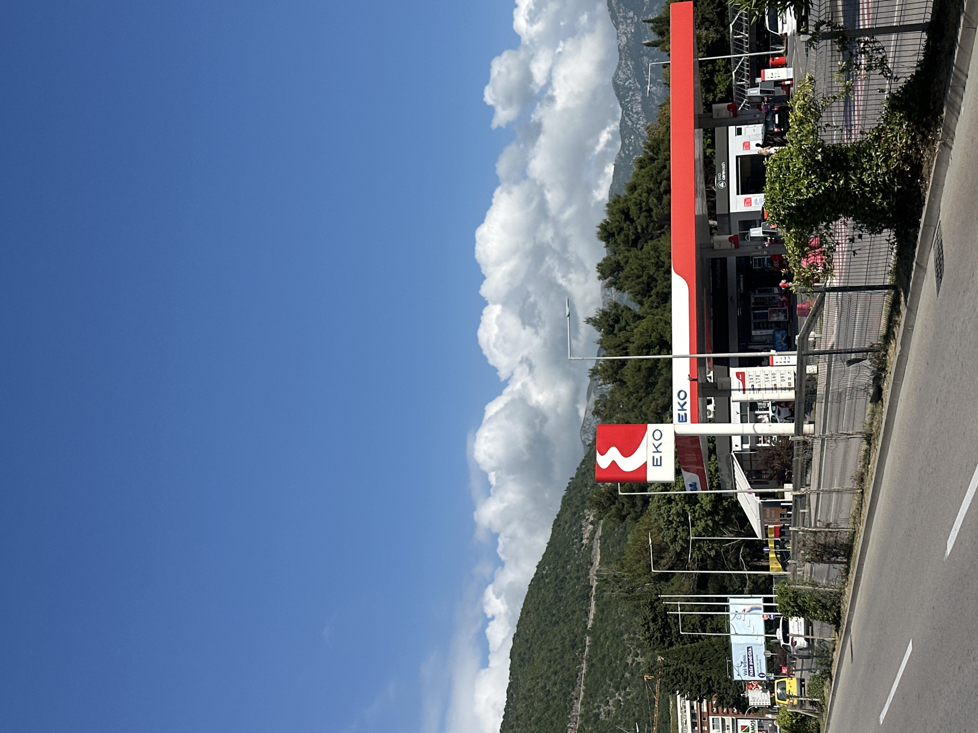 A sideways photograph captures a gas station with its prominent red and white EKO signage set against a backdrop of a bright blue sky and mountains. A large, fluffy cloud formation divides the sky from the mountain range. The gas station, situated on the right side of the frame, is bordered by a road with white lane markings. A wire fence separates the gas station area from the road. Trees and greenery are interspersed throughout the scene. The gas station's red canopy contrasts with the white pumps and signage, creating a striking visual. There's a hint of other buildings and structures at the bottom of the frame, suggesting the presence of a city nearby. This is likely a gas station located near Budva, Montenegro, given the mountain landscape and architecture.