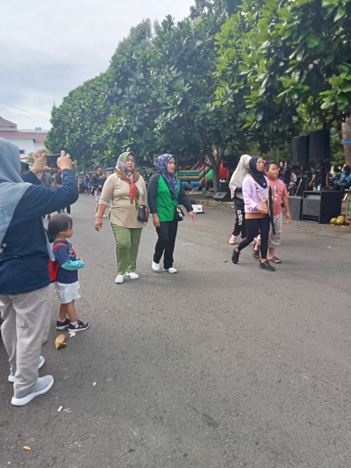 The image captures an outdoor scene in Singaparna, Indonesia, featuring several people on a paved road, likely during a community event on an overcast day.
In the foreground on the left, a person wearing a grey hijab and a dark blue long-sleeved shirt is holding a smartphone horizontally, seemingly recording or photographing something to the right. Next to them, a young child with a red backpack and a light-colored skirt stands, looking towards the right. A discarded banana peel lies on the asphalt near the child's feet.
Further into the scene, two women wearing hijabs walk side-by-side. The woman on the left wears a light green hijab, a colorful patterned scarf, a light-colored top, and olive green pants, carrying a black shoulder bag. The woman beside her has a blue patterned hijab, a bright green long-sleeved shirt with the word "WALK" partially visible, and black pants. Both women wear white shoes.
Behind them, a group includes two younger individuals and a boy. One girl wears a light purple hoodie and black leggings with "ADIDAS" written on the leg. The boy next to her wears an orange t-shirt and light shorts.
The background features lush green trees lining the road. On the right side, large black speakers suggest a stage or performance area, with a colorful inflatable structure visible behind them, indicating a family-friendly event. Distant buildings with white and red roofs can be seen on the left. The overall atmosphere is casual and relaxed.