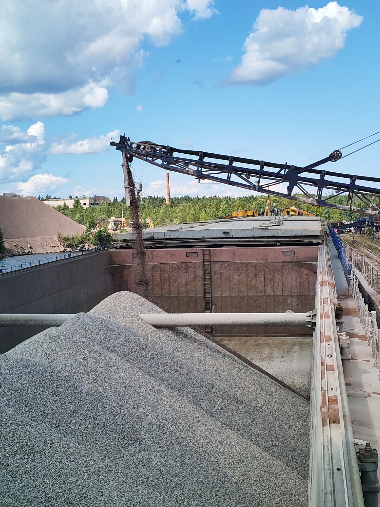 The image depicts an industrial scene in the city of Kvartsitny, Russia, likely at a quarry or processing plant. A large conveyor belt system is actively transferring dark aggregate material from an elevated position into a large, open-topped barge filled with lighter-colored gravel. The barge is positioned alongside a waterway, with a darker brown, rocky embankment visible on the left. In the background, there are large piles of aggregate, industrial structures, trees, and buildings, including a prominent red brick smokestack. The sky is blue with scattered white clouds. The overall impression is one of ongoing material handling and processing in an industrial setting.