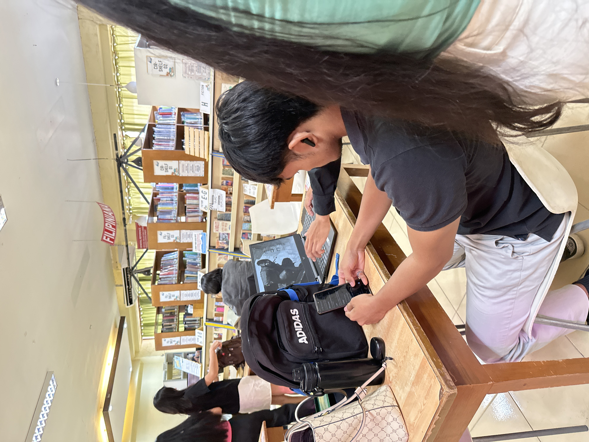 Here is a detailed description of the image:
**Setting:** The image appears to be taken inside a library, as evidenced by the bookshelves filled with books in the background. A sign reading "FILIPINIANA" is visible above some bookshelves, suggesting a section dedicated to Filipino literature or culture. Additional signs are also displayed on the shelves, with tags like "E-G," "H FICTION," "CIR-SHELF 01," and "CIR-SHELF 02." The location is likely in the city of Davao City, Philippines, as indicated by the presence of "FILIPINIANA" sign.
**People:** Several people are present in the image.
* The main subject is a young man wearing a black shirt and light-colored pants. He has short, dark hair and is wearing an earbud. He's holding and looking at a smartphone in his hands.
* Another person is visible partially, typing on a laptop.
* There are also two women in the lower left corner of the image.
**Objects:**
* A black Adidas backpack with a water bottle attached is placed on the table.
* A Gucci purse rests near the backpack.
* A laptop is open on the table, displaying an image on its screen.
* A smartphone is being held by the young man.
**Actions and Interactions:** The young man is using his smartphone, while another person is working on a laptop. The other individuals seem to be occupied as well, likely studying or reading in the library.
**Environment:** The lighting appears to be artificial, provided by ceiling lights. The floor is tiled. The tables are made of wood. Overall, the environment seems to be a typical library setting.