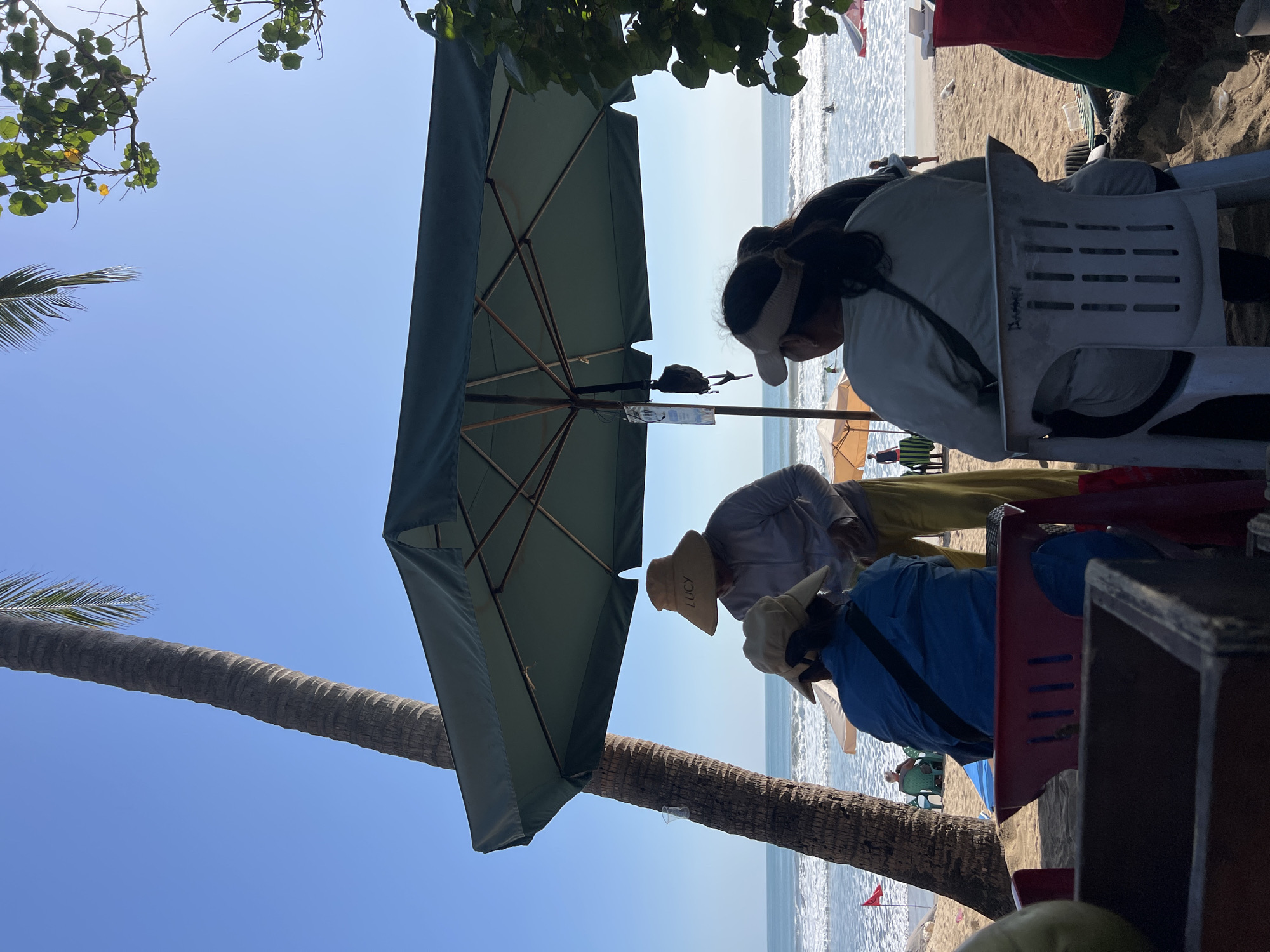This image depicts a beach scene in Kuta, Indonesia, under a clear blue sky. In the foreground, two individuals are seated under a large green umbrella, facing the ocean. The person on the left is wearing a blue shirt and a straw hat, while the person on the right is wearing a light-colored visor and a grey top. A person in a grey shirt and yellow pants, also wearing a straw hat with "LUCY" written on it, stands between them, seemingly interacting with them.
Behind the umbrella, the sandy beach stretches towards the ocean, where gentle waves are breaking. Other beachgoers can be seen in the distance, some sitting on chairs or walking along the shore. A tall palm tree is prominently featured to the left of the frame, its trunk leaning slightly towards the sky. Lush green foliage from a tree overhangs the right side of the umbrella. Several beach umbrellas and chairs are scattered on the sand. A red flag is visible on the left side of the beach. The overall atmosphere suggests a relaxed, sunny day at the beach.