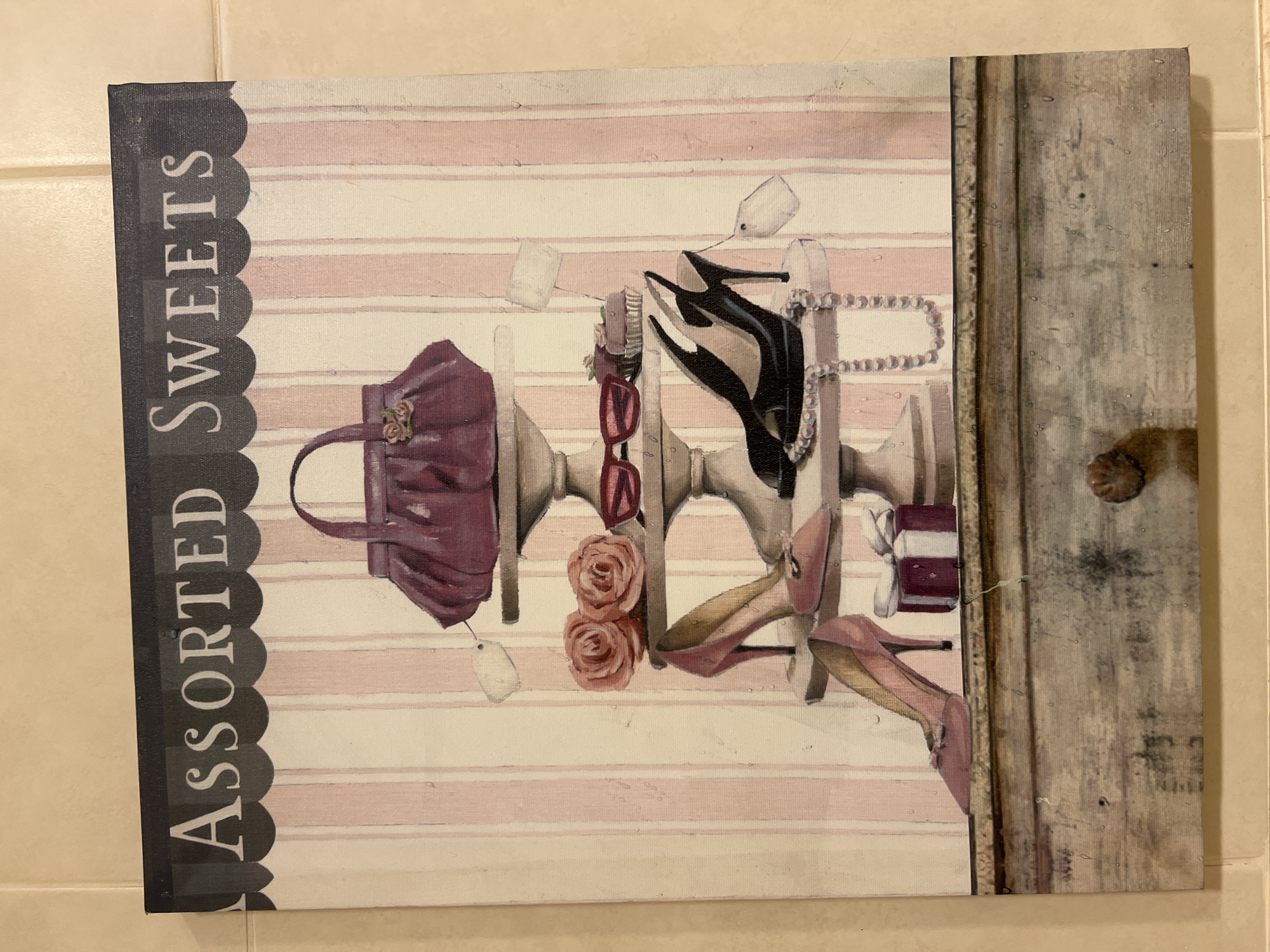 The image is a painting of a tiered display stand with various fashion accessories and small decorative items. The top shelf holds a plum-colored handbag with a floral embellishment. The second shelf features two pink roses and a pair of red-tinted sunglasses. The third shelf displays a pair of pink high-heeled shoes and a small gift box. The bottom shelf has a pair of black high-heeled slingback shoes and a pearl necklace. The background consists of vertical pink and white stripes, topped with a dark border with scalloped edges and the text "ASSORTED SWEETS" in white lettering. Below the display stand is a wooden surface that resembles a drawer front with a knob. The painting is mounted on a wall with beige tiled grout lines visible around the edges. There is no information in the image to suggest a location in Bender, Moldova.
