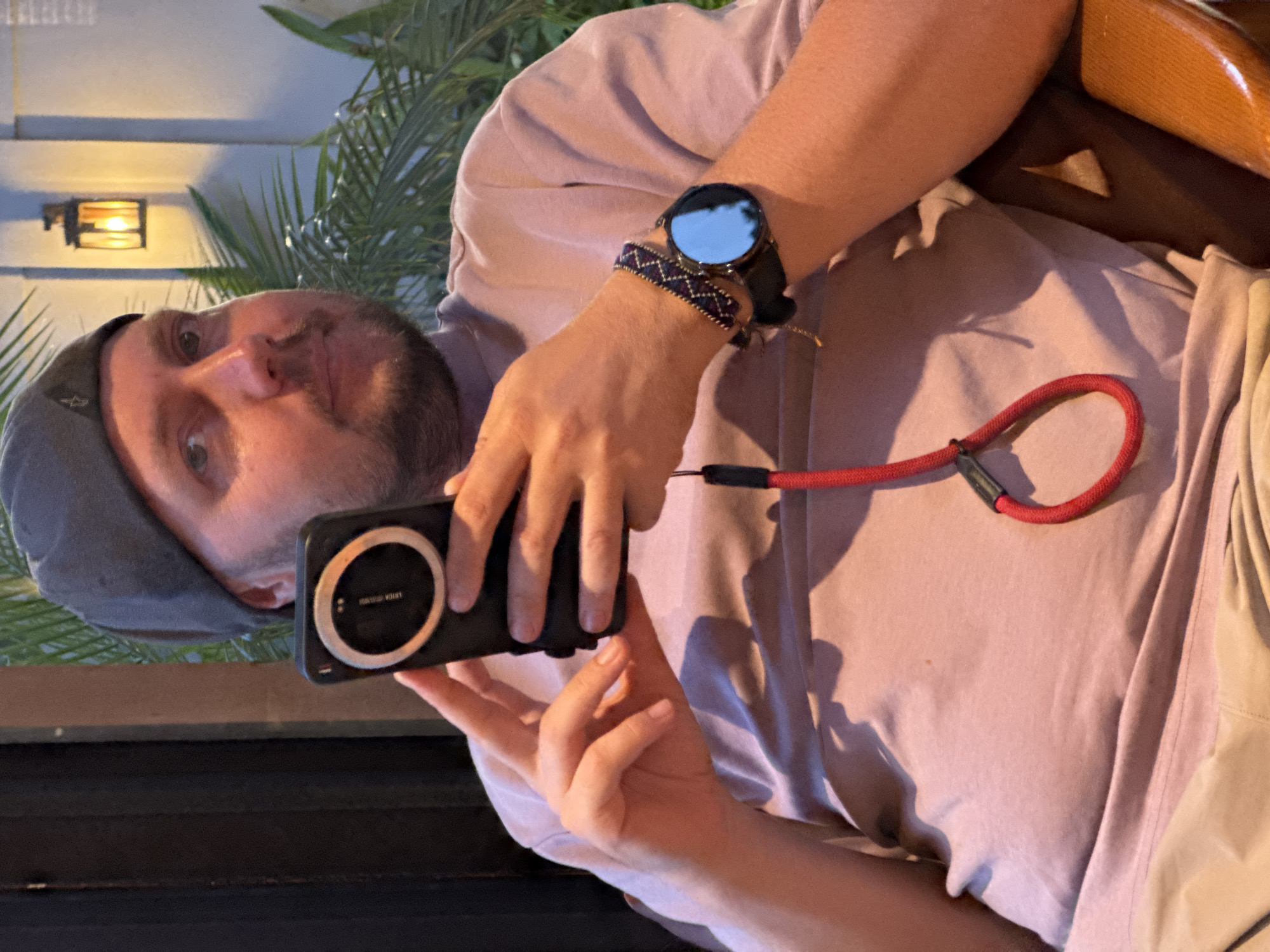 In Nam Hoa Lư Ward, Vietnam, a man wearing a gray beanie and a pink t-shirt is holding a smartphone with a red lanyard. He is also wearing a wristwatch with a black band and a patterned bracelet. The man is sitting in a chair and is looking towards the camera. In the background, there is a wall with a lantern and some palm trees. The lighting is dim, suggesting it is evening or indoors. The man appears to be relaxing or waiting.