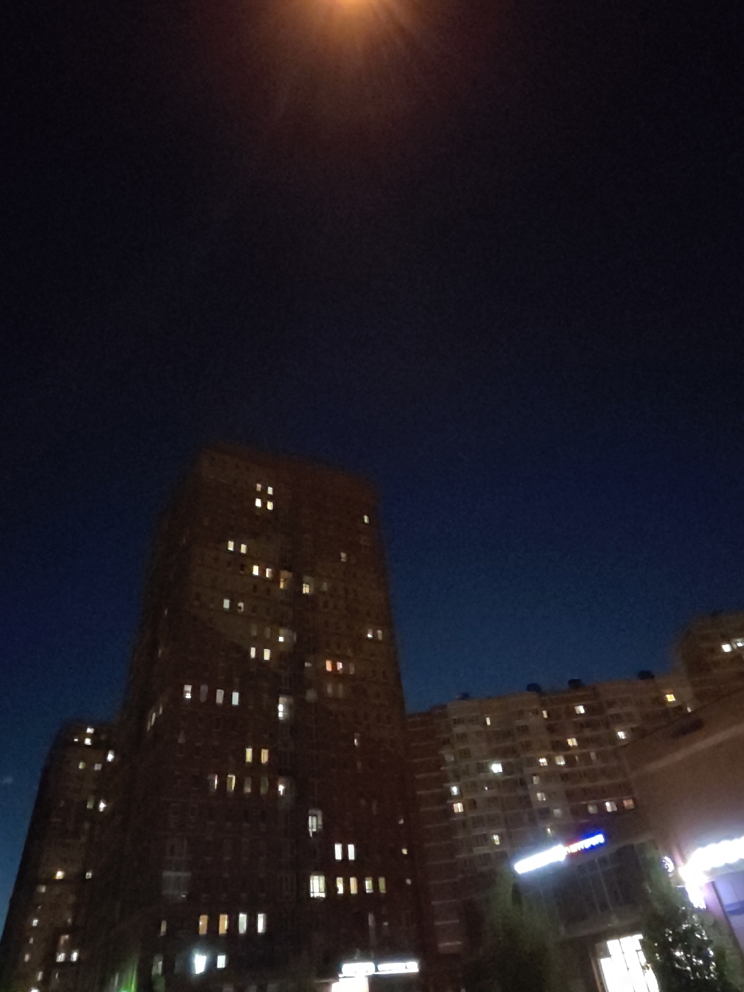 The image depicts a nighttime scene in the city of Shcherbinka, Russia, featuring a dark blue sky with an orange glow from a light source at the top. Two apartment buildings are prominently visible. The building on the left is taller and darker, with many lit windows scattered across its facade. The building on the right is lower and appears to be a commercial area with illuminated signs and windows. The overall impression is of an urban environment at night, with the buildings and lights creating a sense of depth and activity.