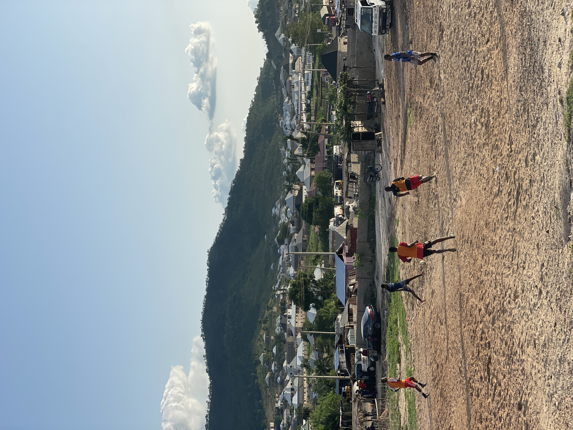 The image shows a view of Lokoja, Nigeria. A mountain covered in vegetation dominates the left side of the frame, with buildings interspersed on its lower slopes. A street runs horizontally across the center of the image, featuring vehicles and motorcycles parked along the side. Several people are on a sandy area adjacent to the street, some of whom appear to be playing soccer. Clouds are visible in the sky.