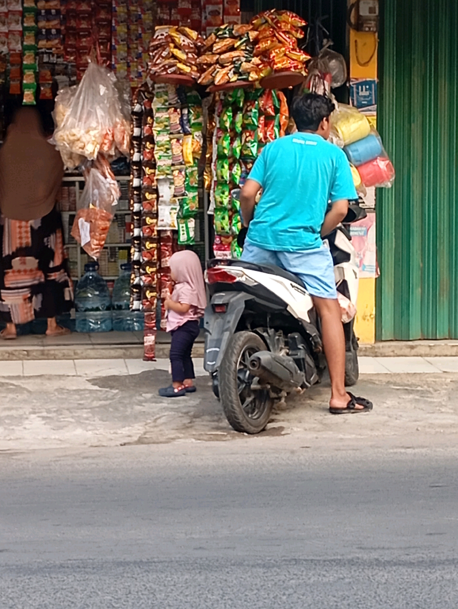 In a street-side store in Babelan, Indonesia, a young child wearing a pink hijab and shirt stands to the right of a parked motorcycle. The child is looking towards the store, which is filled with shelves of snacks and packaged goods. To the left of the child, a person wearing a brown hijab and a patterned dress stands facing the store, partially obscured by a large plastic bag of snacks hanging in front of them. A man wearing a bright blue t-shirt and light blue shorts sits on the motorcycle, facing away from the camera. Behind the store, a green corrugated metal gate is visible. The ground in front of the store is paved with concrete.