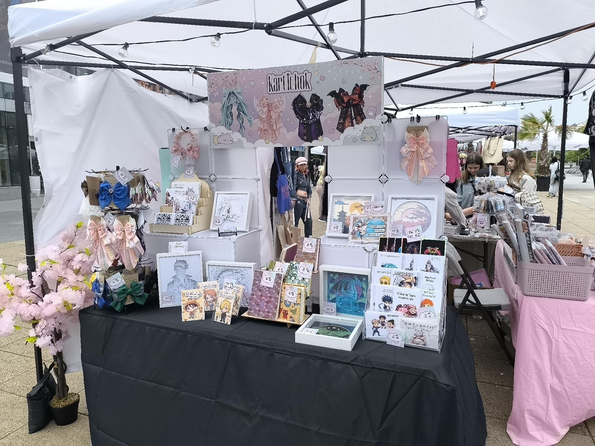 This image captures an outdoor market stall, likely at a craft fair or festival in Burgas, Bulgaria. The stall is covered by a white canopy tent, strung with decorative lights, suggesting a festive atmosphere during daytime. The ground is paved, and palm trees are visible in the background, indicating a warm climate.
The main subject is a merchandise display table covered with a black cloth. A large banner at the back features the name "Kartichok" alongside illustrations of various decorative bows. The stall offers a variety of handmade goods, including numerous hair accessories and bows in different colors (pink, blue, green, black) displayed on stands. Also on sale are art prints, greeting cards, and framed artworks, some appearing to be paper cut-outs or stained-glass style designs, featuring characters from anime/manga like One Piece and Jujutsu Kaisen. An artificial pink cherry blossom tree decorates the left side of the display.
Two young women, customers, are seen browsing items on the right side of the stall. Another person in a pink cap is visible further back. Various price tags are visible, showing amounts such as "2€", "8€", "15€", "17€", "20€", "22€", and "25€". Other visible text includes "THE PSYCHO PRINCESS", "THE POTATO PRINCE", "OPEN ME", "Magic exists / it makes / people happy", "Happy Crocs Birthday!", "I'll keep my fingers / crossed for you", "You are / the best!", "お誕生日おめでとう" (Happy Birthday in Japanese), "BEST BRO", and "I believe in you".