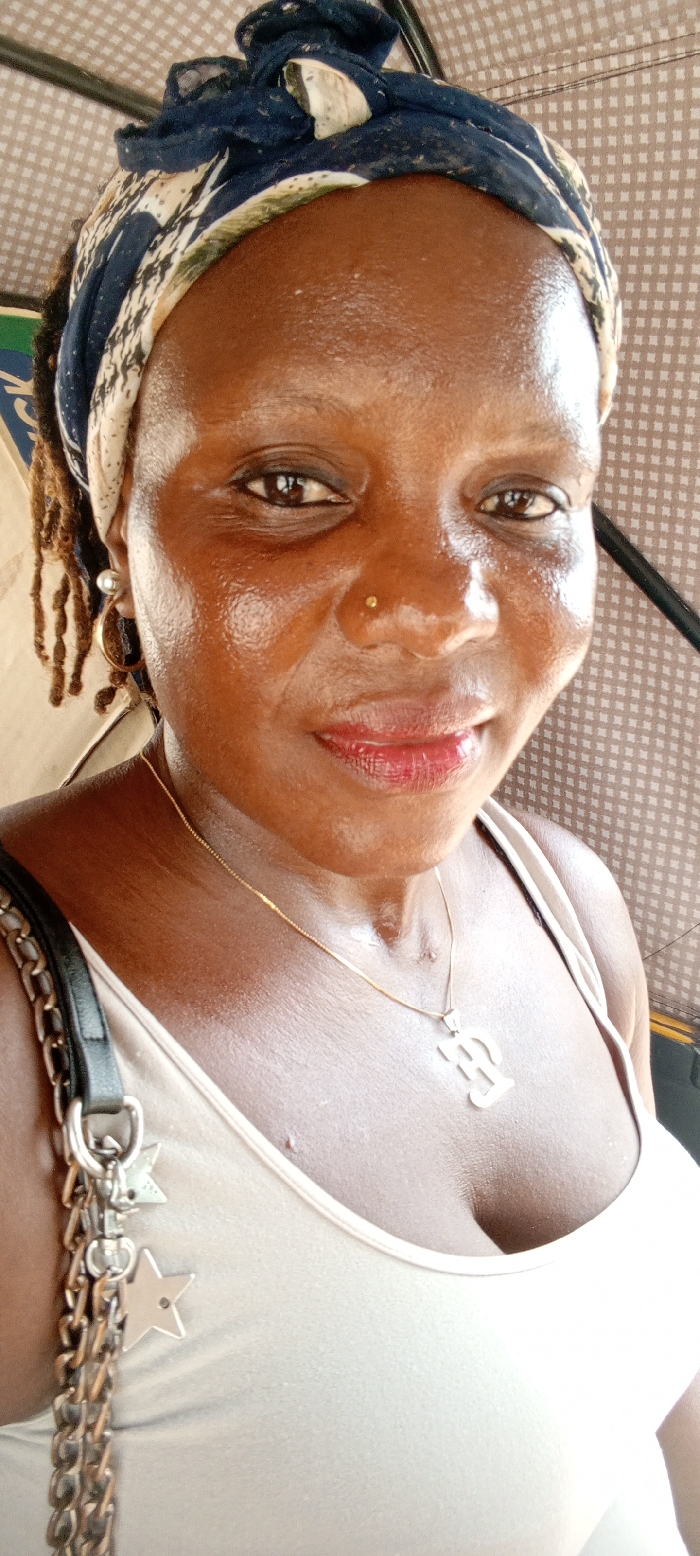 A close-up portrait features an adult female with medium-dark skin, looking directly at the camera with a slight smile. She wears a dark blue headwrap patterned with white and lighter elements over what appears to be dreadlocks, partially visible on her left. Her right nostril bears a small gold-toned stud. Her eyes are dark brown with visible eyeliner, and her lips are painted with a reddish-berry hue. A thin gold-toned chain with a pendant resembling the letters "EE" or "EF" is around her neck, resting on a light-colored tank top. A black bag strap with a silver-toned chain and two star-shaped charms hangs over her left shoulder. The background consists of a light brown/beige checkered fabric, consistent with the interior roof lining of a vehicle. The scene is located in the city of Iranla, Nigeria.