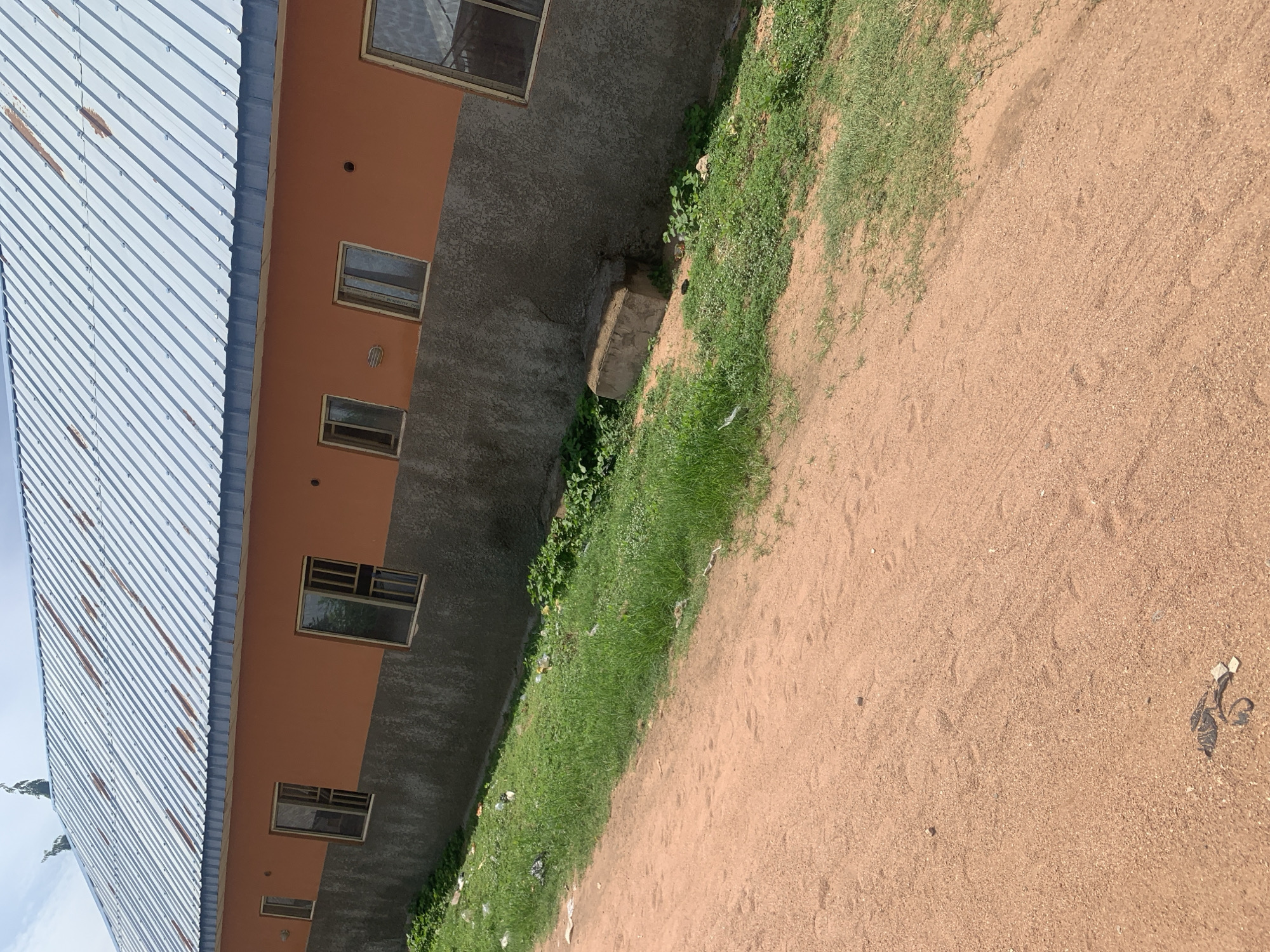 The image shows a side view of a building with an orange upper wall and a gray lower wall, topped with a corrugated metal roof. The roof has some rust spots. There are several windows on the upper wall, most with metal bars. The building is situated on a grassy incline that slopes down to a dirt road in the foreground. Scattered on the grass and the dirt road are pieces of debris, including plastic bags and paper. The environment appears to be outdoors with a clear sky. This scene is located in Jauro Yinu, Nigeria.