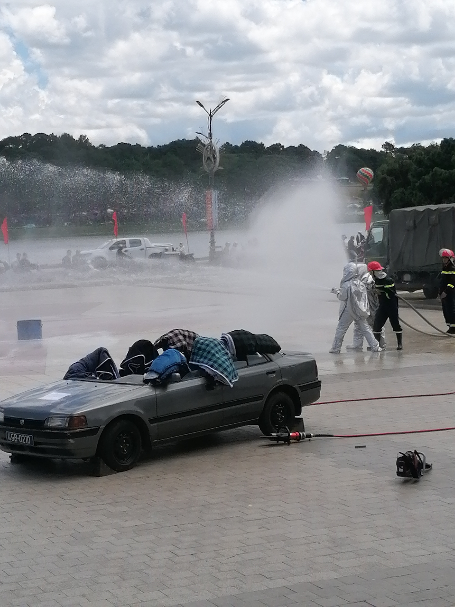 The image captures an outdoor scene in Da Lat, Vietnam, during what appears to be a fire rescue drill or demonstration. In the foreground, a gray sedan with a license plate reading "LSB-0210" is parked, with various checkered and solid-colored cloths draped over its interior. A hydraulic rescue tool, often called the "jaws of life," is positioned next to the car, connected by a red hose.
Further in the mid-ground, several individuals in protective suits, including one in a silver hazmat suit and others in firefighting gear, are engaged in an activity involving a large water spray. One person in the silver suit is holding a hose, directing the water spray. Behind them, a military-style truck is parked. In the background, a white pickup truck is partially submerged in water, with people riding on it and around it, suggesting a simulated rescue scenario. A hot air balloon is visible in the sky, and trees line the horizon. The ground is paved with light-colored interlocking bricks. The sky is overcast with dramatic clouds.