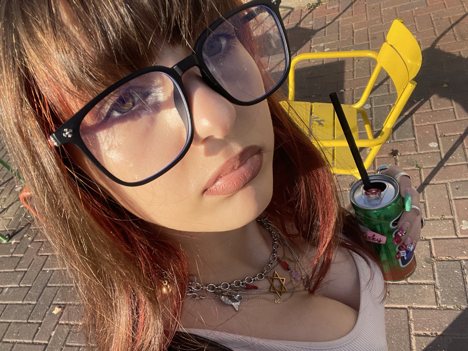 An overhead photograph shows a person from the chest up, positioned against a brick-paved ground and a yellow metal structure. The subject has reddish-brown hair with lighter highlights, styled to frame the face. Black-framed glasses with silver cross-like embellishments on the temples cover the eyes; the left eye, visible through the lens, appears light brown. The person's lips are painted with a matte, light reddish-brown lipstick. Around the neck are multiple silver-toned chain necklaces adorned with various charms, including a prominent golden Star of David, a small silver-colored fish, and several red teardrop-shaped pendants. The left hand is visible, holding a green aluminum can with a black straw inserted into its opening. The can features graphic designs and a sticker with cursive writing, possibly "lucky." The fingernails are decorated with multicolored polish, and multiple silver rings are worn on the fingers. The background consists of reddish-brown brick pavers and a bright yellow metal frame, possibly part of a playground or public seating, casting distinct shadows under direct sunlight. The specific location within Raanana, Israel, cannot be determined from the visual information provided.