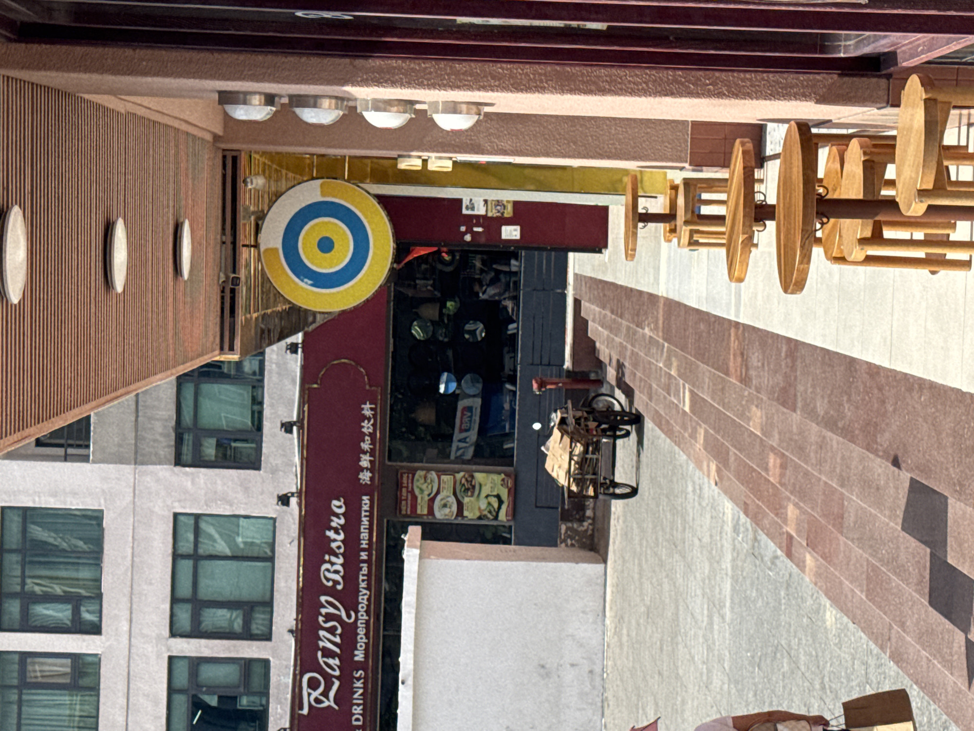 The image depicts a street scene in Bac Nha Trang Ward, Vietnam. In the foreground, a row of small wooden tables and stools are set up outside a building, suggesting an outdoor seating area for a cafe or restaurant. The tables are round, and the stools have simple wooden bases. The building has a pinkish-brown textured facade with vertical dark brown panels on the right side, possibly part of a shopfront. Above the tables, a wooden ceiling with horizontal slats is visible, with round lights embedded in it.
Further back, a sign with stylized script reads "Pansy Bistro," indicating the establishment's name. Below this, text in English, Russian, and Chinese characters suggests a diverse clientele or offerings. To the left of the bistro's entrance, a white rectangular structure stands. The entrance itself is recessed and appears to lead into a dimly lit interior. A circular sign with concentric blue and yellow rings is mounted above the entrance.
In the middle ground, a three-wheeled cart loaded with cardboard boxes is positioned on the paved walkway. The cart has two large rear wheels and a smaller front wheel, and appears to be either parked or in the process of being moved. A red fire hydrant is visible next to the cart. The ground surface is a mix of grey paving stones and reddish-brown tiled steps or pathways.
In the background, a multi-story building with many windows is visible. The windows have dark frames and appear to be covered with light-colored curtains. The overall impression is a commercial area in Vietnam, with a mix of modern and traditional architectural elements.