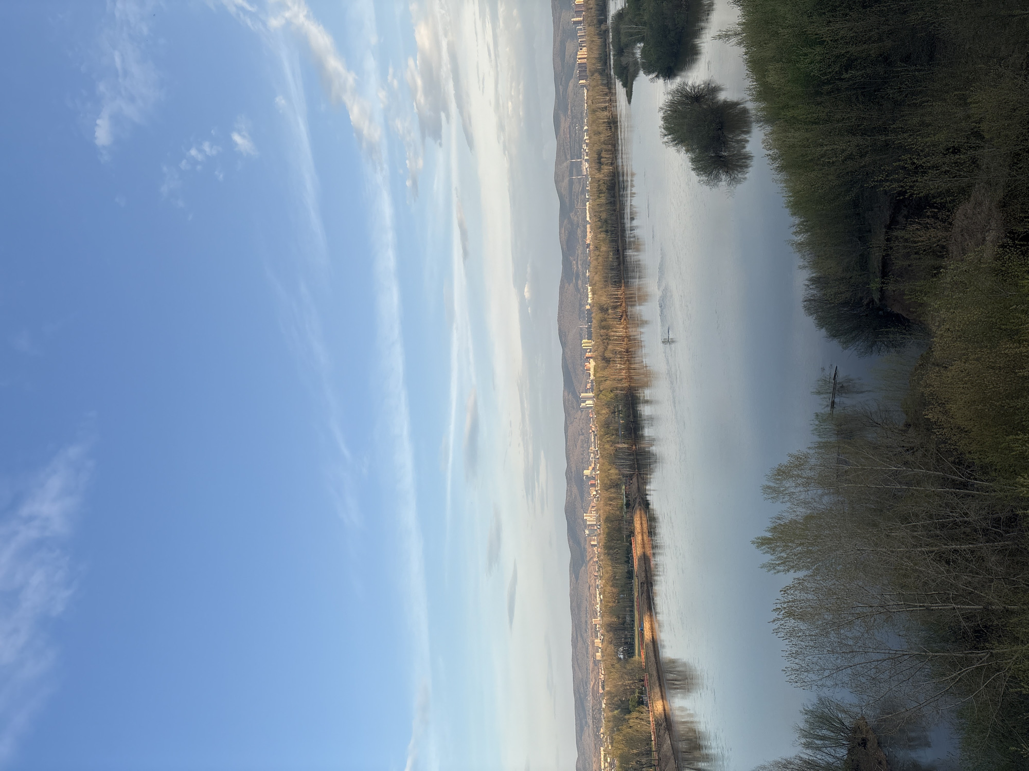 The image shows a river with trees on the right bank. In the background, there is a city against a backdrop of mountains. The left side of the image shows a blue sky with elongated clouds. The city is Unknown, Unknown.