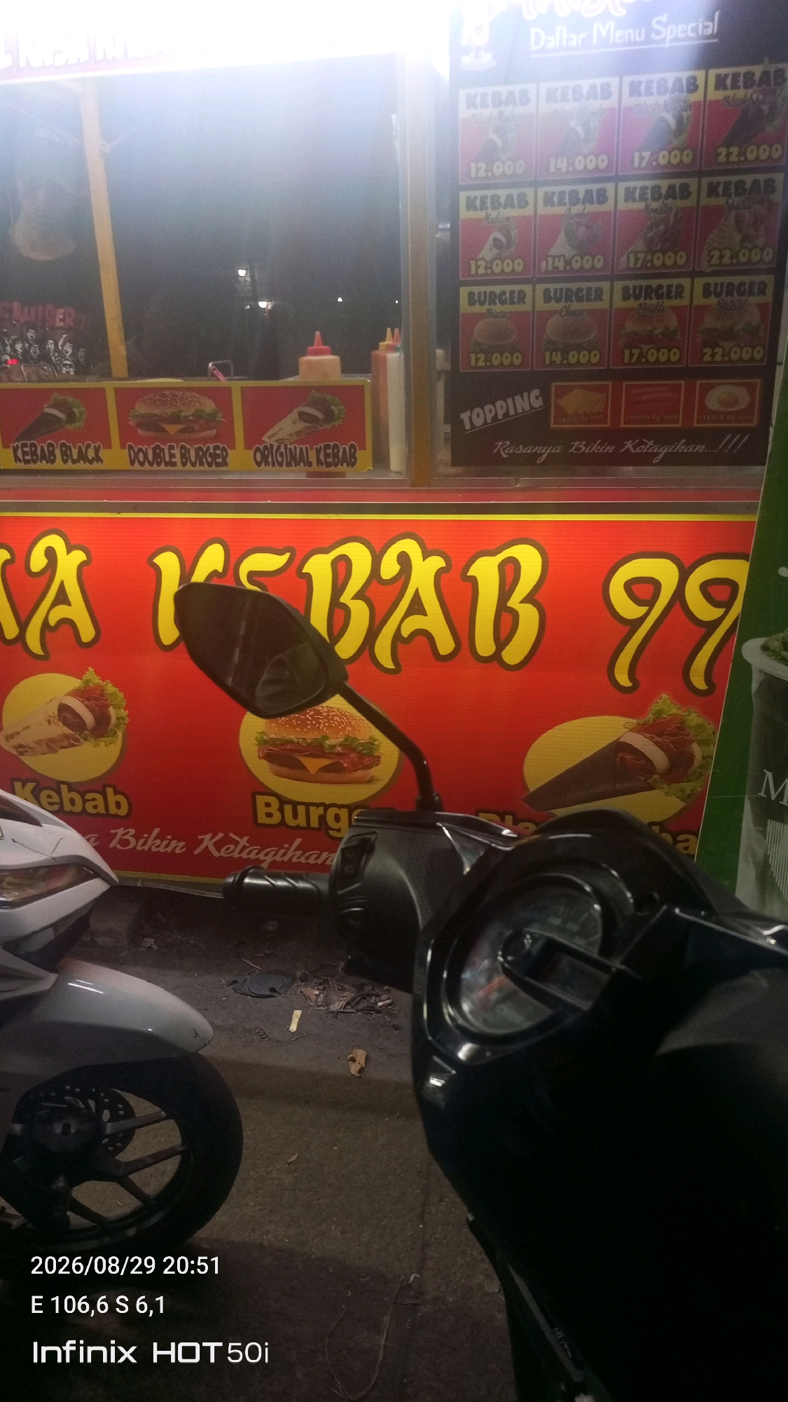 The image captures a street-side food stall at night in Kampung Melayu Barat, Indonesia. The stall features a bright red banner with yellow lettering that reads "MA KABAB 99", indicating it sells kebabs and possibly burgers. Various menu items are displayed on the stall's facade, including images and prices for different types of kebabs and burgers. Condiment bottles, likely ketchup and chili sauce, are visible. In the foreground, parts of two motorcycles are present, suggesting the location is a popular spot for commuters or locals. The overall lighting suggests an outdoor, urban setting at dusk or night.