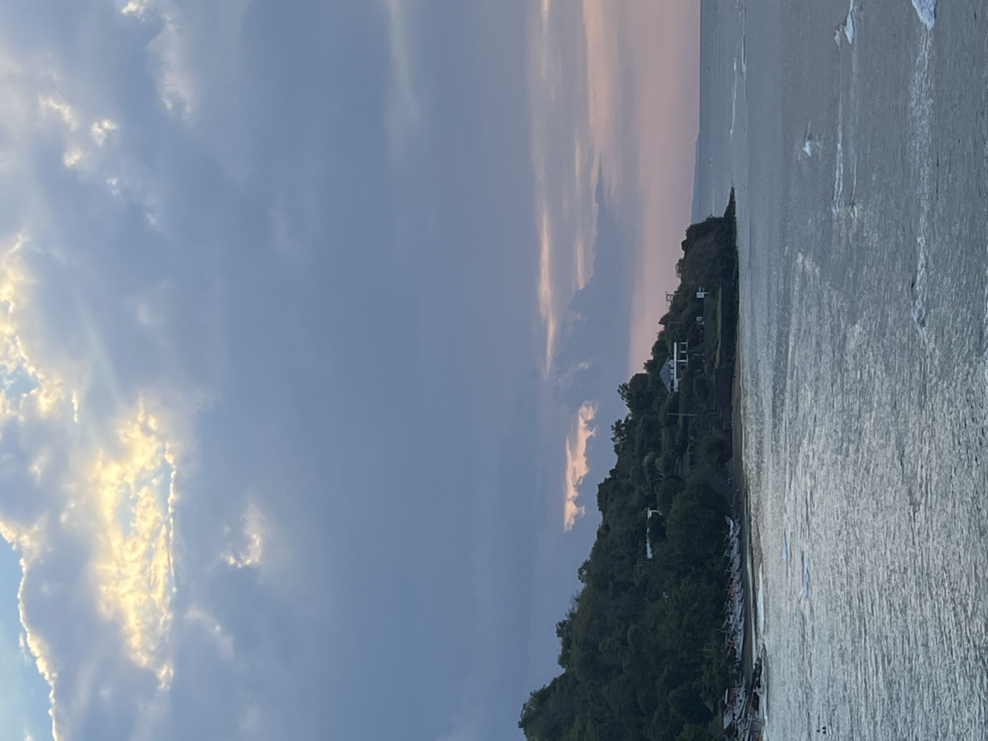 The image depicts a scenic view of a coastline at dusk, likely in Abang, Indonesia. The sky is a canvas of dramatic clouds, with patches of sunlight breaking through, casting a warm glow against the softer hues of pink and lavender near the horizon. Below the sky, a lush, green hillside slopes down towards the sea. A few buildings are visible nestled among the trees on the hillside, suggesting a residential or resort area. Along the shore, several small fishing boats are pulled up onto the sand, lined up in a row. The sea itself is a deep, textured blue, with gentle waves creating white foam as they approach the shore. The overall atmosphere is one of tranquility and natural beauty, characteristic of a tropical coastal evening.