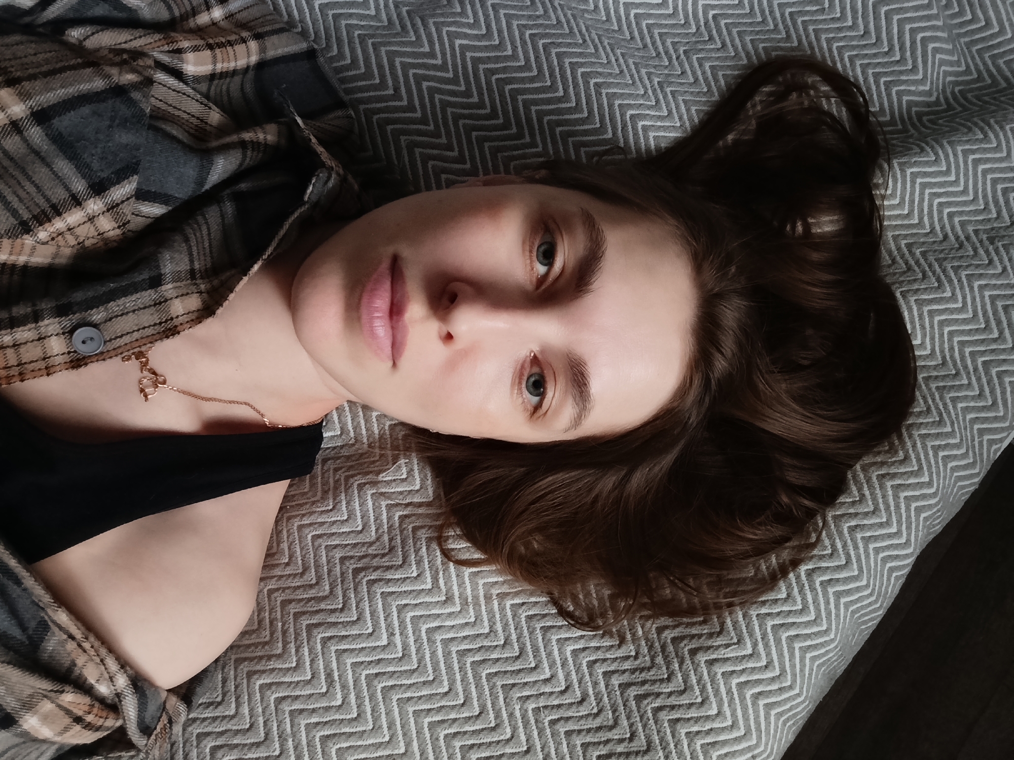 A young woman lies on a gray and white chevron patterned surface. She is wearing a black top underneath a tan, gray, and brown plaid flannel shirt, with a gold necklace. Her dark brown hair is spread out on the surface behind her head. She is looking at the camera. The location cannot be determined from the image.