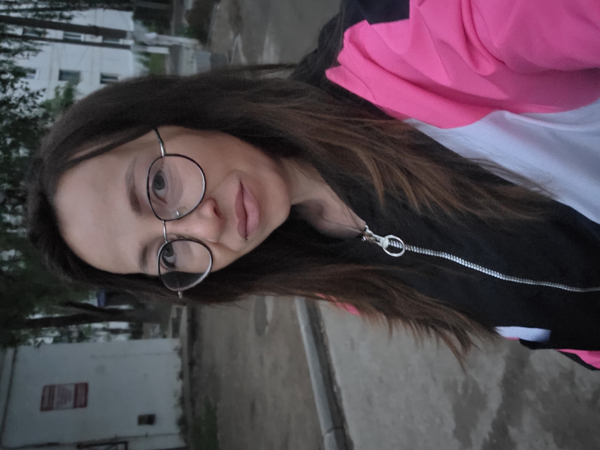 A woman with long, dark brown hair and glasses is standing outside in what appears to be a residential area. She is wearing a black, pink, and white jacket with a zipper. The background shows a white building with a red sign, trees, and a paved area. The overall lighting suggests it is either early morning or late evening. The location is identified as Chaikovsky, Russia.