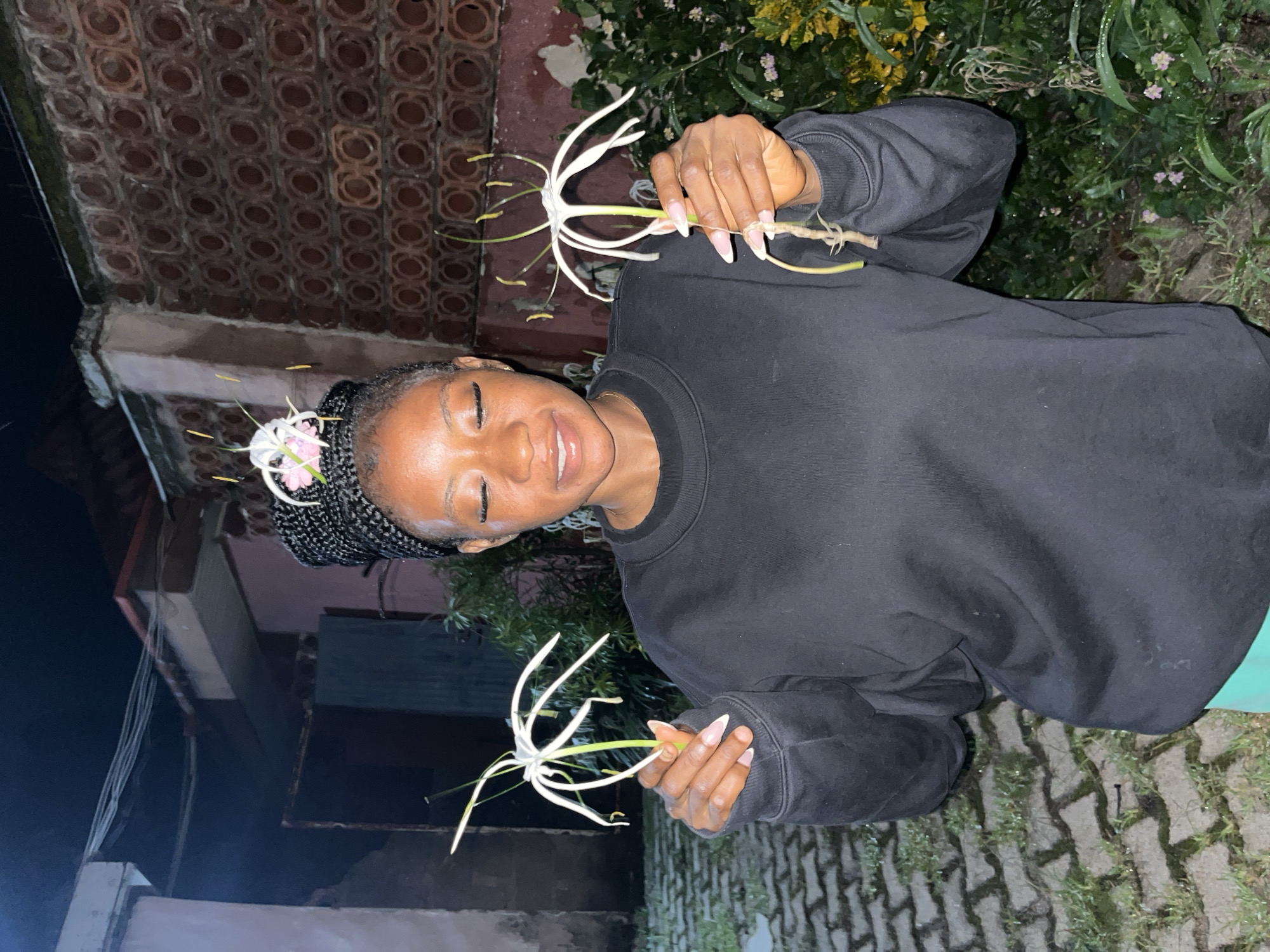 An outdoor medium shot captures a Black woman with her eyes closed, smiling slightly, as she holds two delicate white spider lilies. One flower is positioned in front of her chest, held by her right hand, with its delicate petals fanned out. The other flower is held by her left hand, positioned slightly to her left, also displaying its intricate bloom. The woman has her hair braided in cornrows, with a few of the white petals of the spider lily woven into her hairstyle. She is wearing a black crewneck sweatshirt and appears to be outdoors at night, indicated by the dim lighting and the dark sky. The background features a textured, patterned brick wall and some foliage. The ground is paved with interlocking bricks, and there is evidence of recent rain or dew on the ground and plants. The overall atmosphere is serene and artistic, suggesting a peaceful moment in nature or a decorative arrangement. Based on the provided context, the image was taken in Jos, Nigeria.
