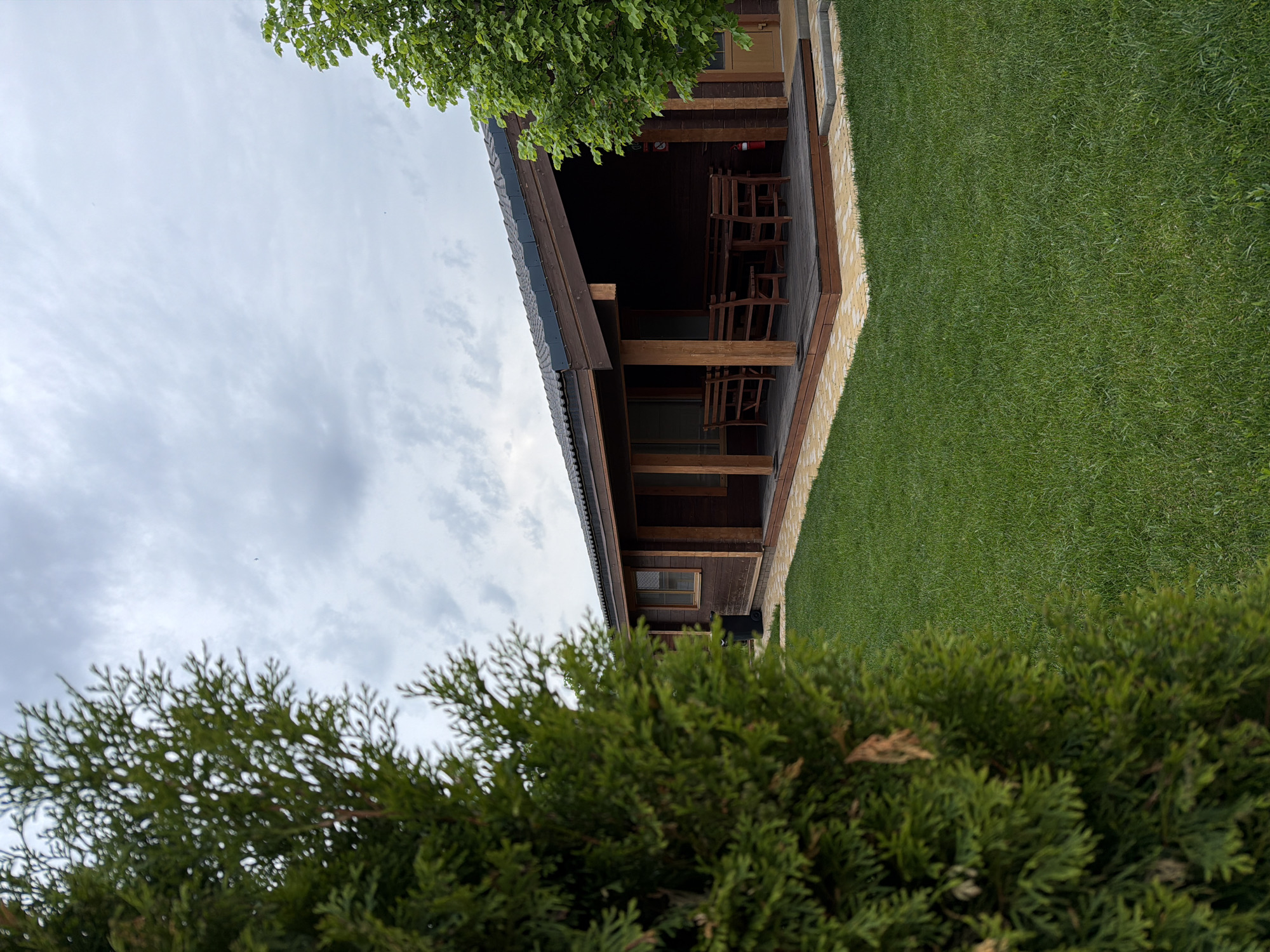 The image depicts a single-story wooden building in a green, grassy environment, likely taken in a rural or suburban area. The building has a covered porch area furnished with wooden chairs and what appears to be a fire extinguisher mounted on the wall. The sky is overcast. A coniferous shrub occupies the foreground. The image does not provide explicit evidence of its location in Kamyshin, Russia. Without additional visual markers or location tags, confirmation is impossible.