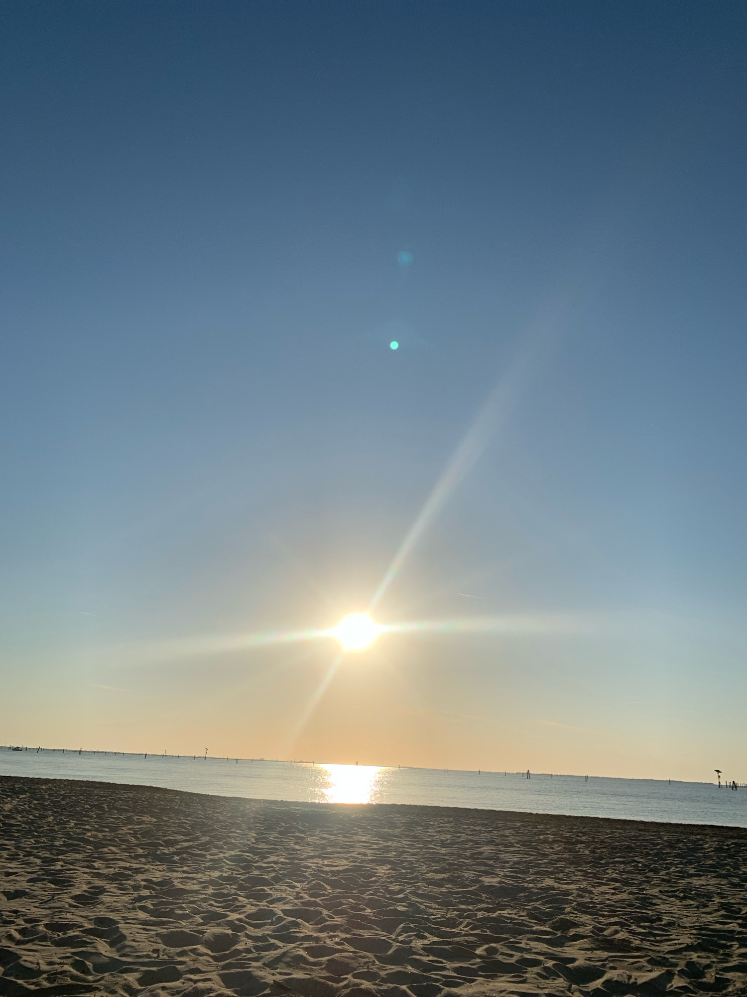 A low-angle, vertical, full shot captures a beach at what appears to be sunset or sunrise. The foreground is dominated by dark, rippled sand, with footprints visible. The middle ground features a calm body of water, likely the sea, with a faint line of buoys or markers stretching across the horizon. In the distance, a single small boat is visible on the left side of the frame, and on the right side, a dark structure resembling a small shack or shelter stands on stilts. The sky is a clear, pale blue, transitioning to a warmer yellow-orange hue near the horizon. The sun is a bright, starburst-shaped object, positioned in the upper-middle of the frame, casting strong lens flares and light rays across the sky and water. Two faint, circular lens flares are also present in the upper portion of the sky. The overall scene suggests a serene and tranquil beach environment. This scene is likely located in the Po Delta region of Comacchio, Italy, known for its extensive wetlands, lagoons, and coastline.