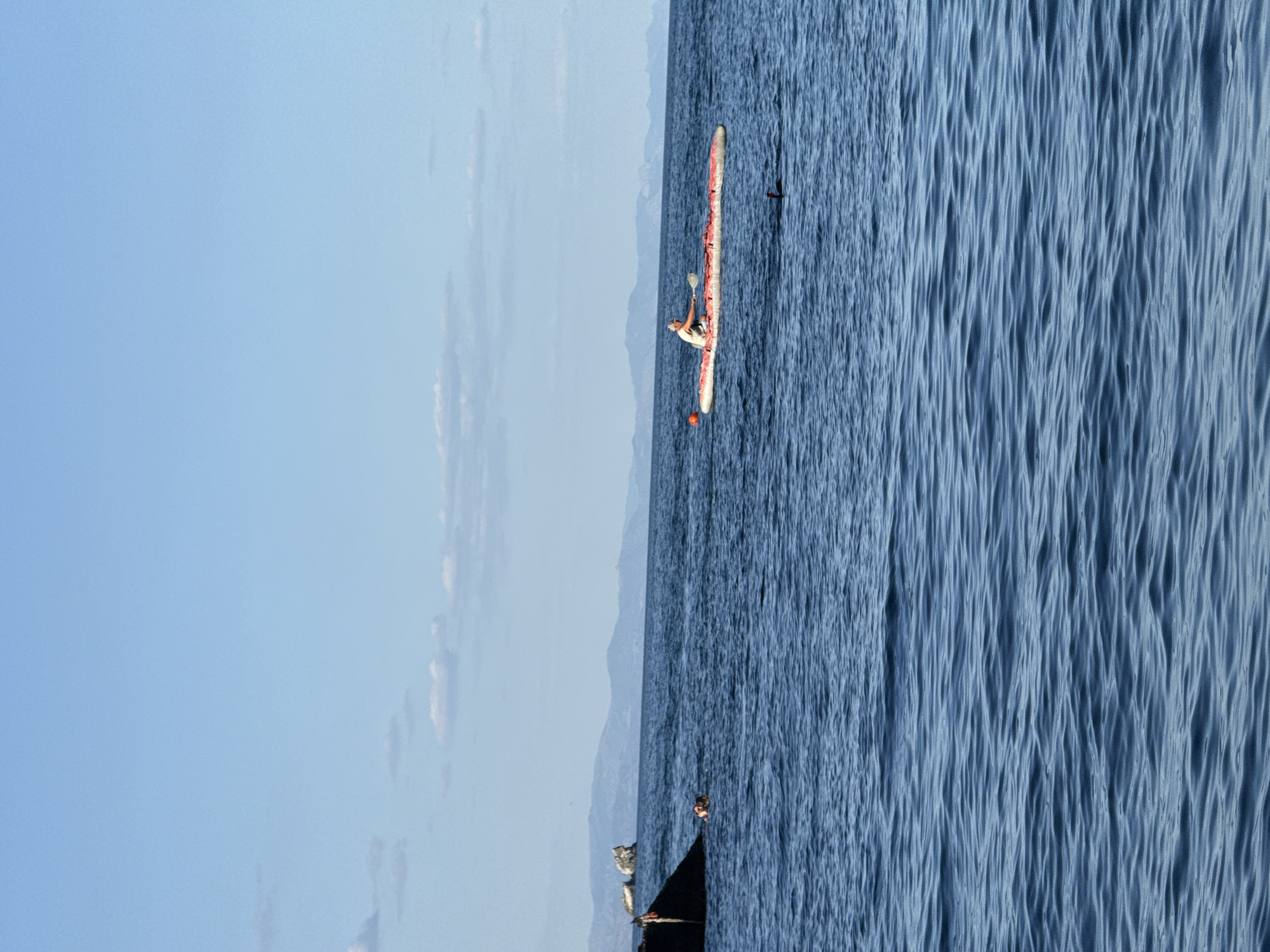 In the foreground, deep blue rippling water fills the lower half of the frame. In the middle ground, a person is paddling a long, red and white kayak across the calm sea. To the left, a dark, angular structure juts out from the water, and near it, another person appears to be swimming. In the background, hazy blue mountains line the horizon under a clear, light blue sky with scattered white clouds. The overall scene suggests a recreational activity on a sunny day.