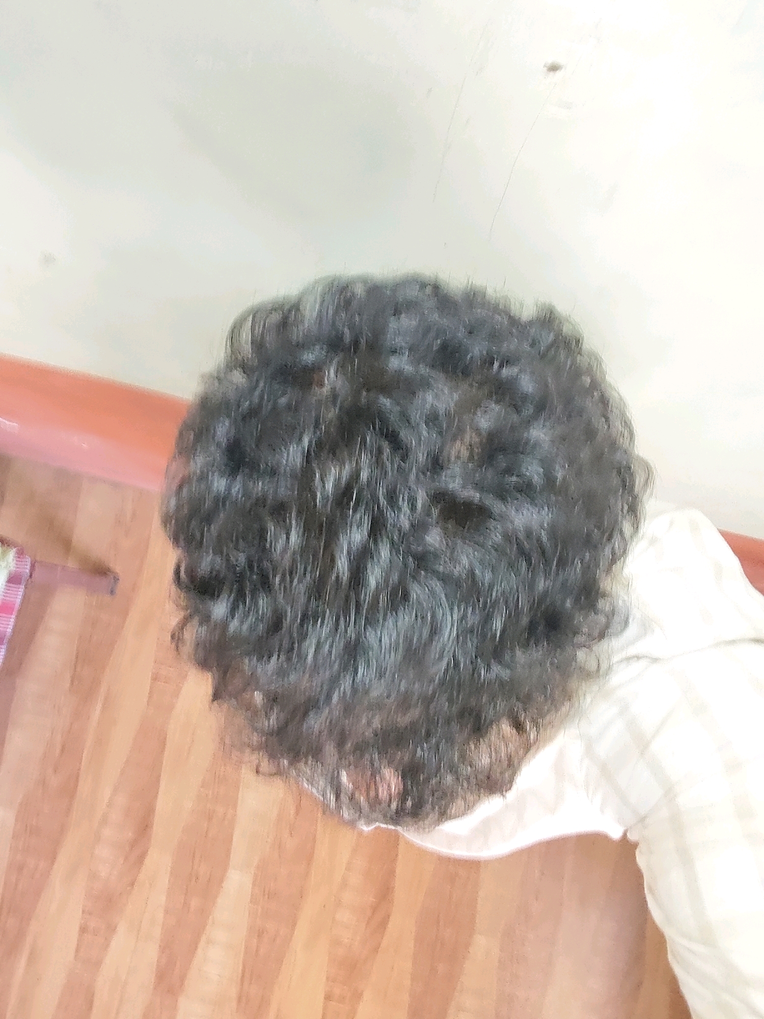 The image shows the back of a person's head, covered in dark, curly hair. The person is wearing a light-colored, checkered shirt. They are positioned on what appears to be a wooden floor with distinct planks. To the left, there is a hint of a red and white checkered object, possibly a mat or rug, and an orange-colored horizontal surface, likely a wall or furniture. The background wall is a pale, off-white color. There is a small dark speck on the wall, potentially a bug or dust. The lighting is soft and diffused, creating some shadows and highlights on the hair and floor. The scene is indoors, and given the detail of the wooden flooring and the general aesthetic, it could be a residential setting within Belagavi, India.