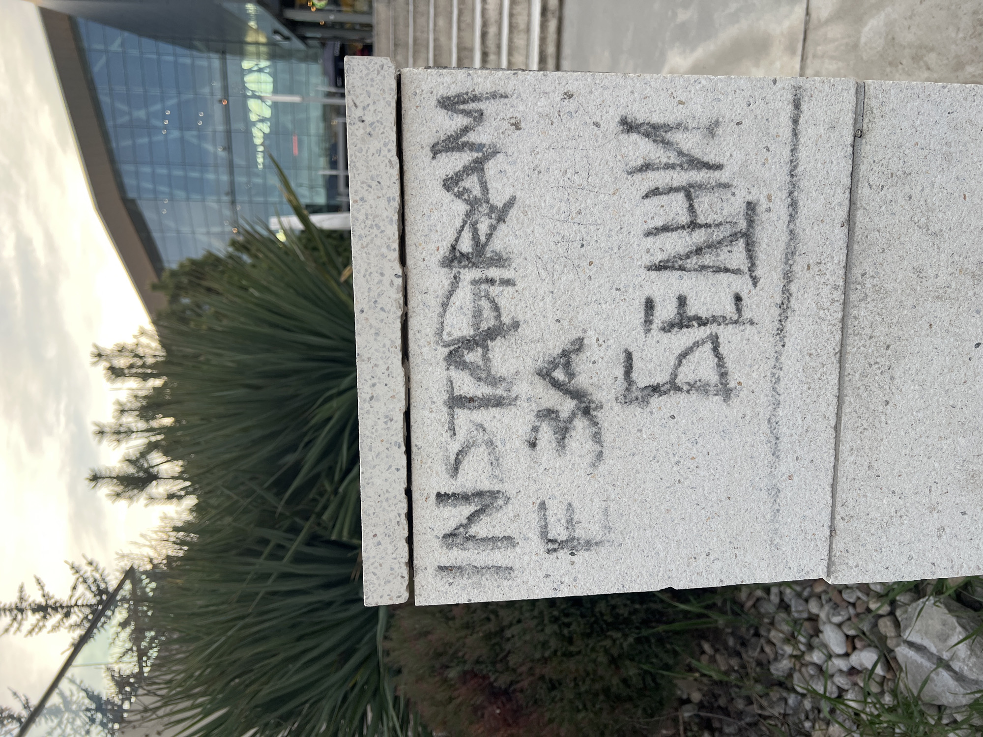 A close-up of a light-colored concrete pillar with graffiti on it. The graffiti says "INSTAGRAM" and then what appears to be a username, which is difficult to fully decipher but might be "IHN3A FEAT.UA". The pillar is in front of some greenery and pebbles, with a modern glass building and stairs visible in the background. The photo appears to have been taken outdoors. The picture was probably taken in Sofia, Bulgaria.