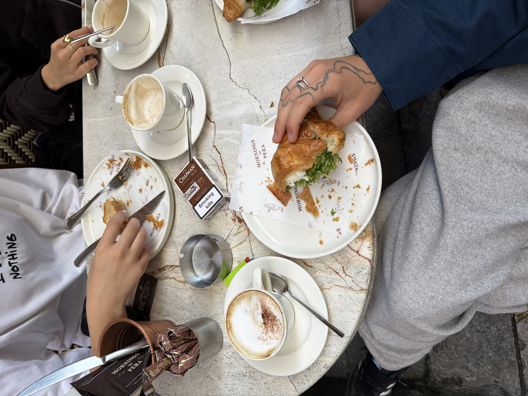 A high-angle, eye-level shot depicts a café table scene. Two people sit at the table with partially consumed meals and drinks. The person on the left wears a white sweatshirt with the word "NOTHING" printed on it. Their hands are holding cutlery and there is croissant on the plate. The person on the right wears a navy blue long-sleeved top and grey sweatpants; their tattooed hand holds half of a croissant sandwich. The table is marble with two empty coffee cups and saucers, one full cup of cappuccino and saucer, two white plates containing remnants of food and croissants, cutlery, an ashtray, a container holding sugar packets, and a pack of Chapman No.4 cigarettes. The packaging of the croissants and the sugar packets indicate the café is "Mornings in Pera" located in Beyoğlu, Turkey.