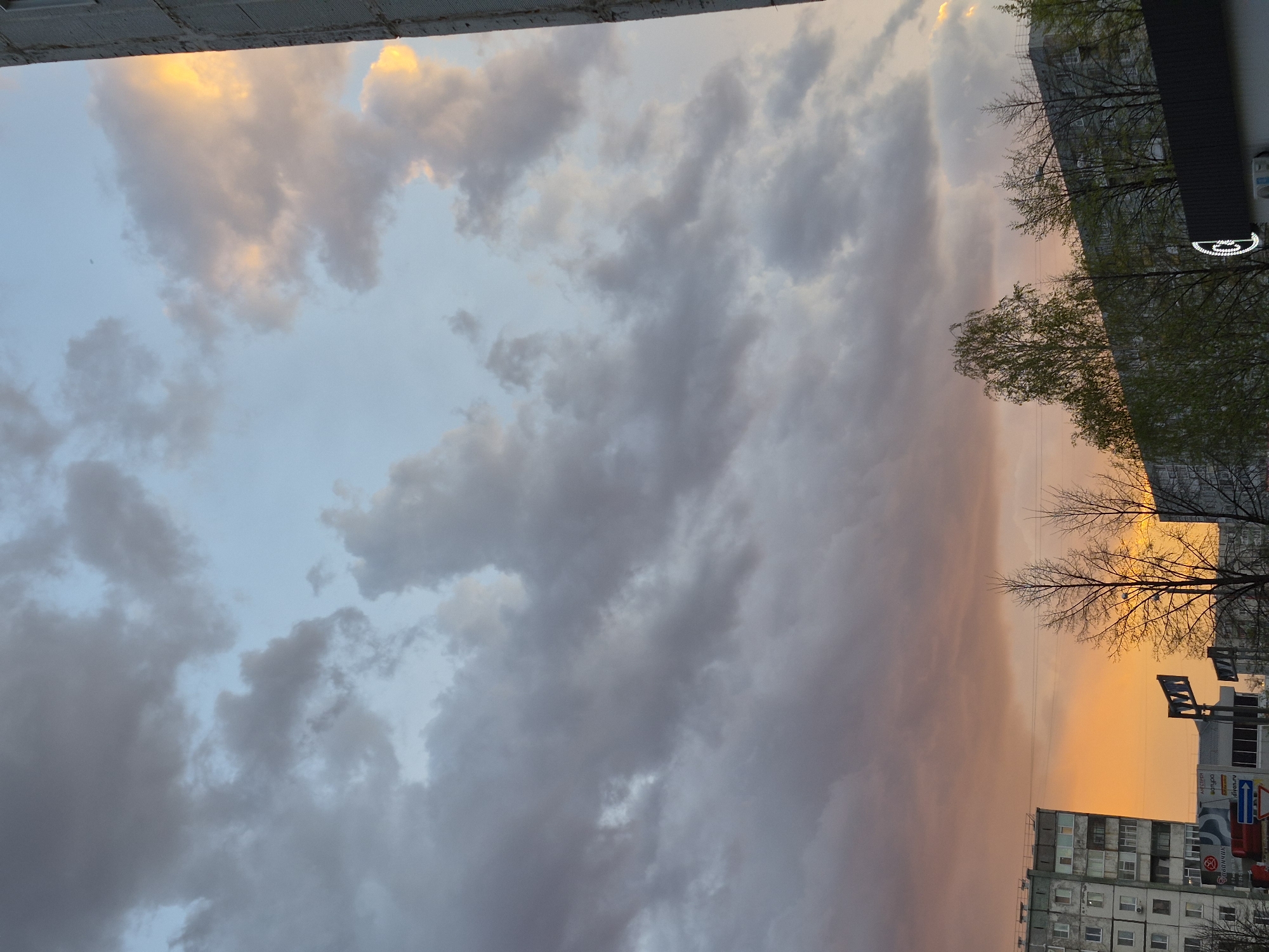 The image depicts a cityscape at sunset, with a predominantly cloudy sky transitioning from blue to orange. The clouds are a mix of gray and white, illuminated by the setting sun. Below the clouds, there are several multi-story residential buildings, typical of urban architecture. A few bare trees are visible in the foreground, partially obscuring the buildings. In the lower right corner, there is a street sign and commercial signage, suggesting a populated area. The overall environment suggests a city at dusk. The location is Tobolsk, Russia.