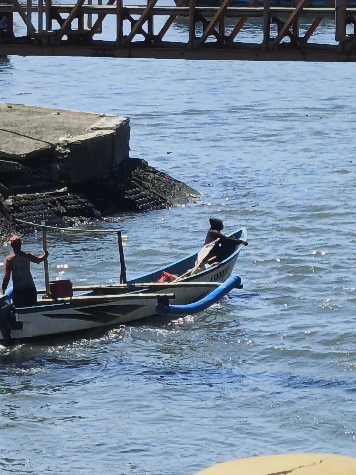 Two people are in a boat on the water. One person is standing at the stern of the boat, wearing a red cap and a red-sleeved shirt. This person appears to be holding onto a wooden pole. The second person is seated in the bow of the boat, wearing a dark hooded garment. The boat is white with a dark stripe and has a blue tube attached to its side. In the background, a concrete structure, possibly a pier or dock, is visible along the water's edge, with some debris on it. A metallic structure, likely a bridge or walkway, is in the upper portion of the image, spanning over the water. The water is blue and choppy, with sunlight reflecting off its surface. The location is identified as Kelurahan Tegalkamulyan, Cilacap, Indonesia.