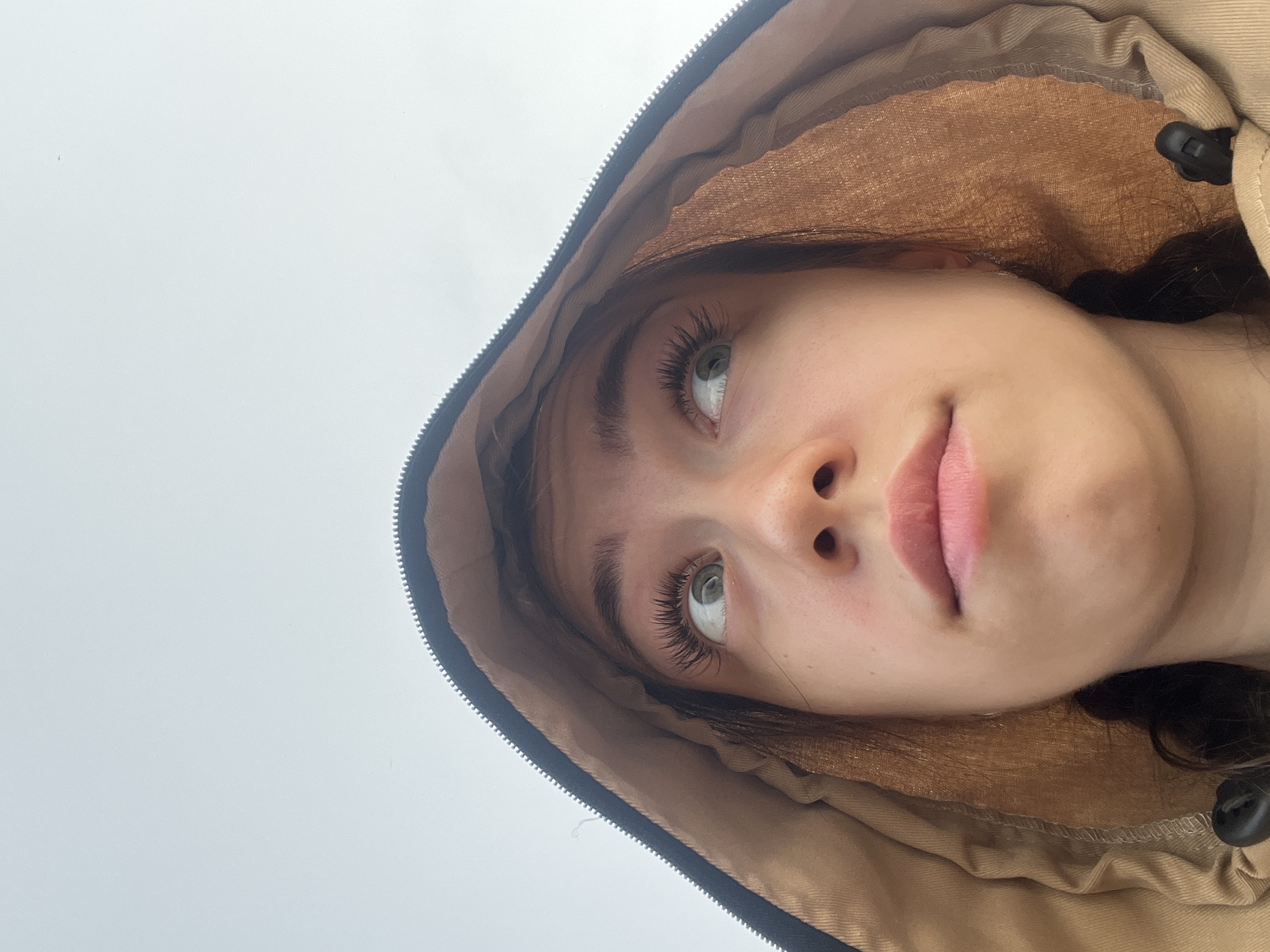 This is a close-up, low-angle, outdoor, natural light, eye-level shot of a young person's face, looking upwards and to the right. The person is wearing a hooded jacket, which frames their face. The background is a pale, overcast sky. The person has light brown hair styled in two braids. Their eyes are green and their eyelashes are long and dark. Their lips are closed and slightly parted. The jacket is tan on the inside and dark on the outside, with a visible zipper. The image appears to have been taken in the city of Chernushka, Russia.