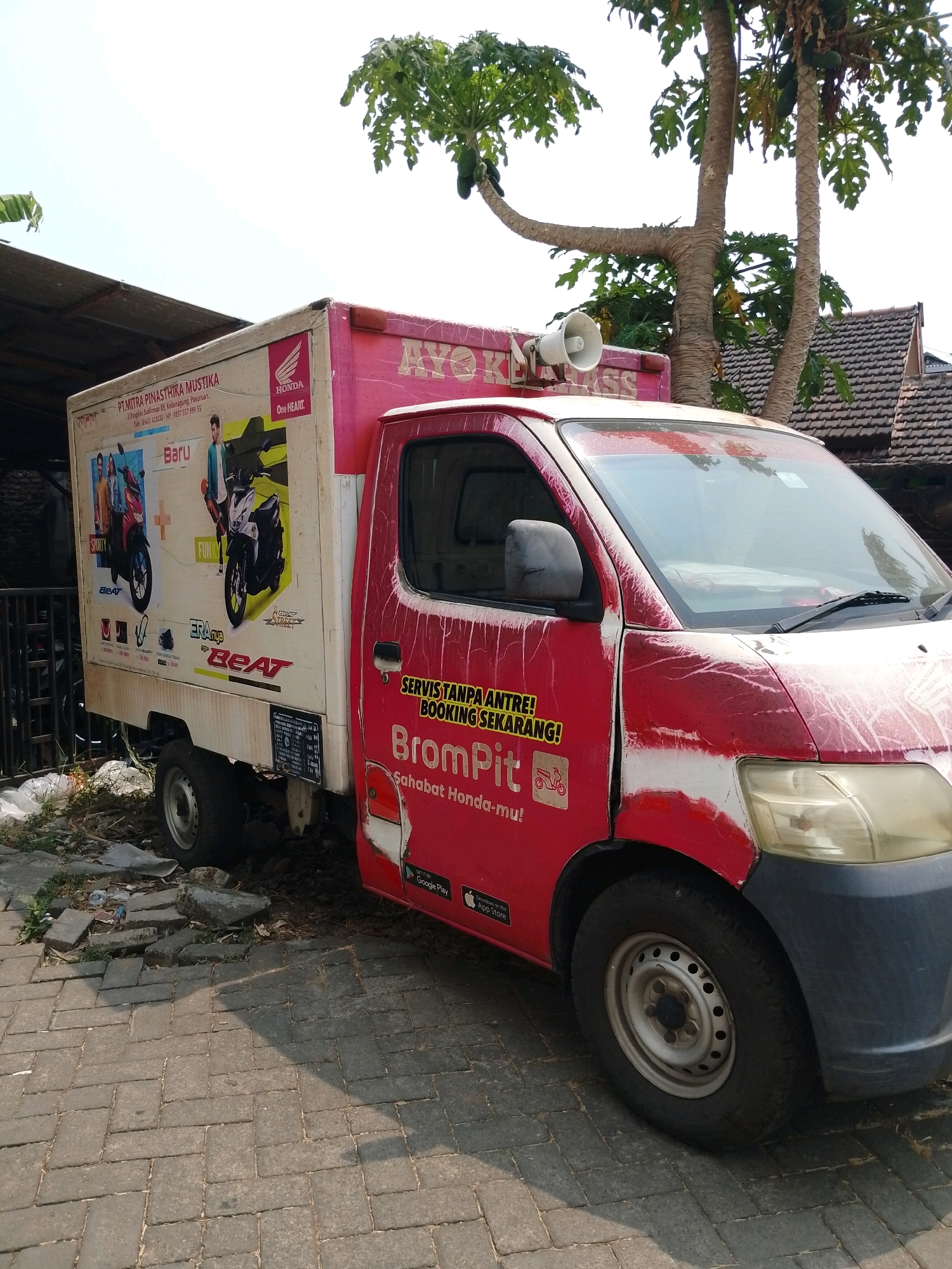 The image shows a red and white delivery truck parked on a brick-paved street in Pasuruan, Indonesia. The truck is branded with "AYO KEMASS" and "BromPit," suggesting it is used for deliveries or transportation services. The truck also features logos for Honda and the "Beat" motorcycle model. The side of the truck has advertisements for "PT. MITRA PINASTHIKA MUSTIKA," a company located at Jl. Anggrek No. 68, Kebonagung, Pasuruan. The vehicle is seen parked near a building with a tiled roof, and lush green trees with visible papaya fruits are growing nearby. The surrounding environment appears to be an outdoor setting, possibly a street or a service area. The truck's paint is weathered, showing signs of wear and tear, with red paint peeling off in several areas, revealing white primer underneath. A megaphone is visible mounted on the top of the truck's cargo box.
