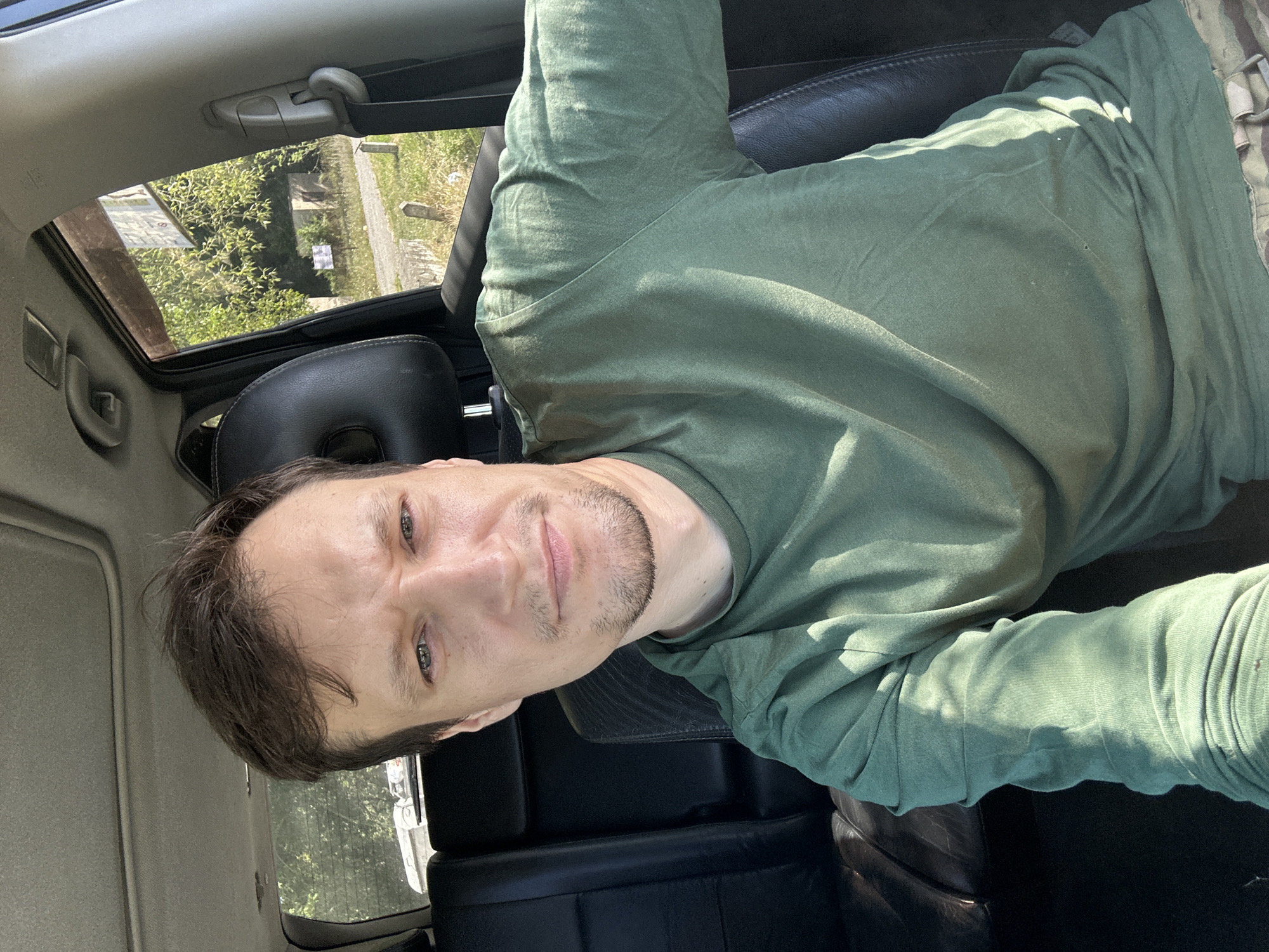 A man is taking a selfie from the driver's seat of a vehicle. He is looking directly at the camera with a neutral expression. He is wearing a green long-sleeved shirt. The interior of the car is visible, including black leather seats and the car's headliner with a sunroof. Through the open passenger window, trees and a dirt road are visible. The location is Makiv, Ukraine. The lighting suggests it is daytime, with sunlight casting shadows on the man's shirt.