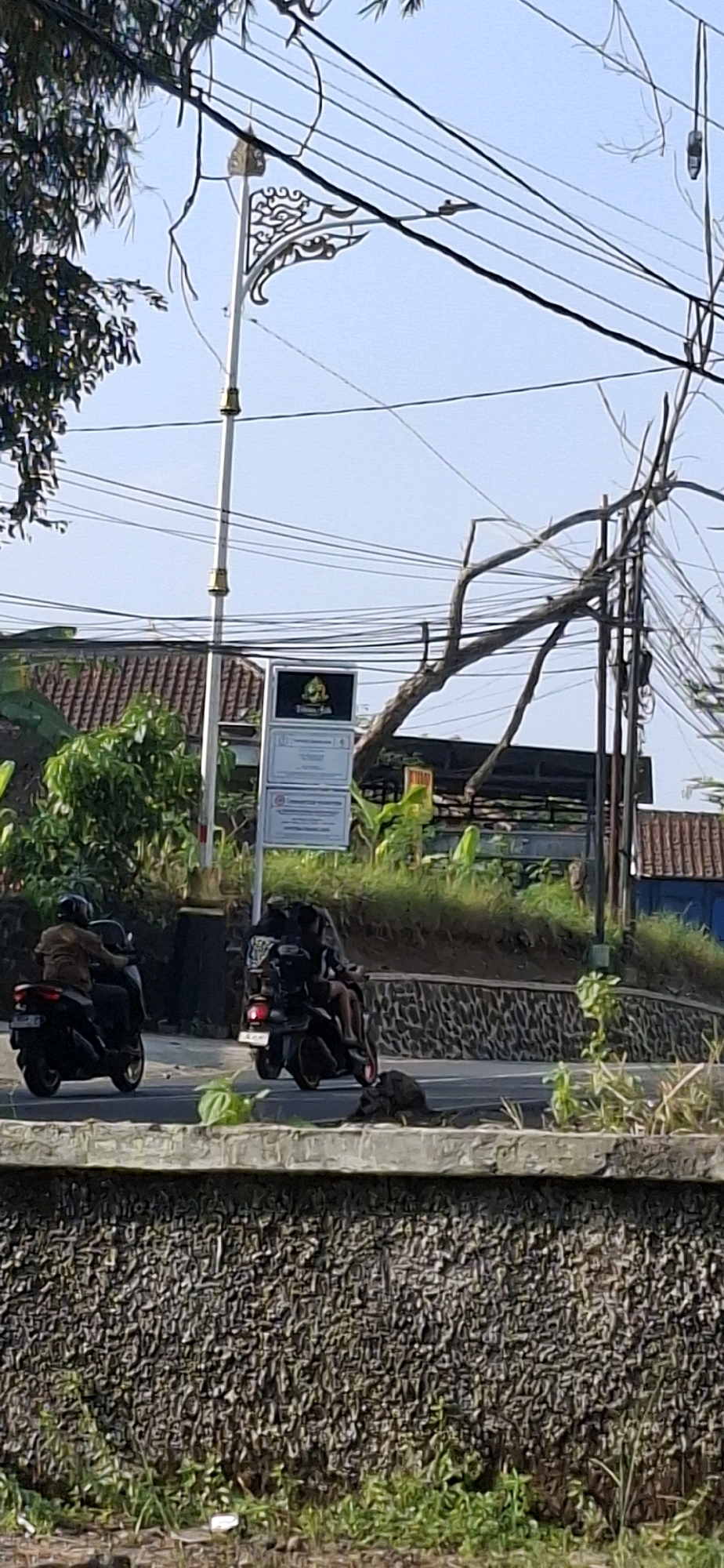In Cilaku, Indonesia, two motorcycles are traveling on a road. The motorcycle on the left is ridden by a person wearing a brown jacket. The motorcycle on the right is carrying two people. Both motorcycles are heading away from the camera. In the background, there are several power lines, a street lamp with ornate decorative elements, and a building with a tiled roof. Green foliage and trees are also visible in the scene. A stone wall with a rough texture runs along the foreground. The sky is clear and blue.